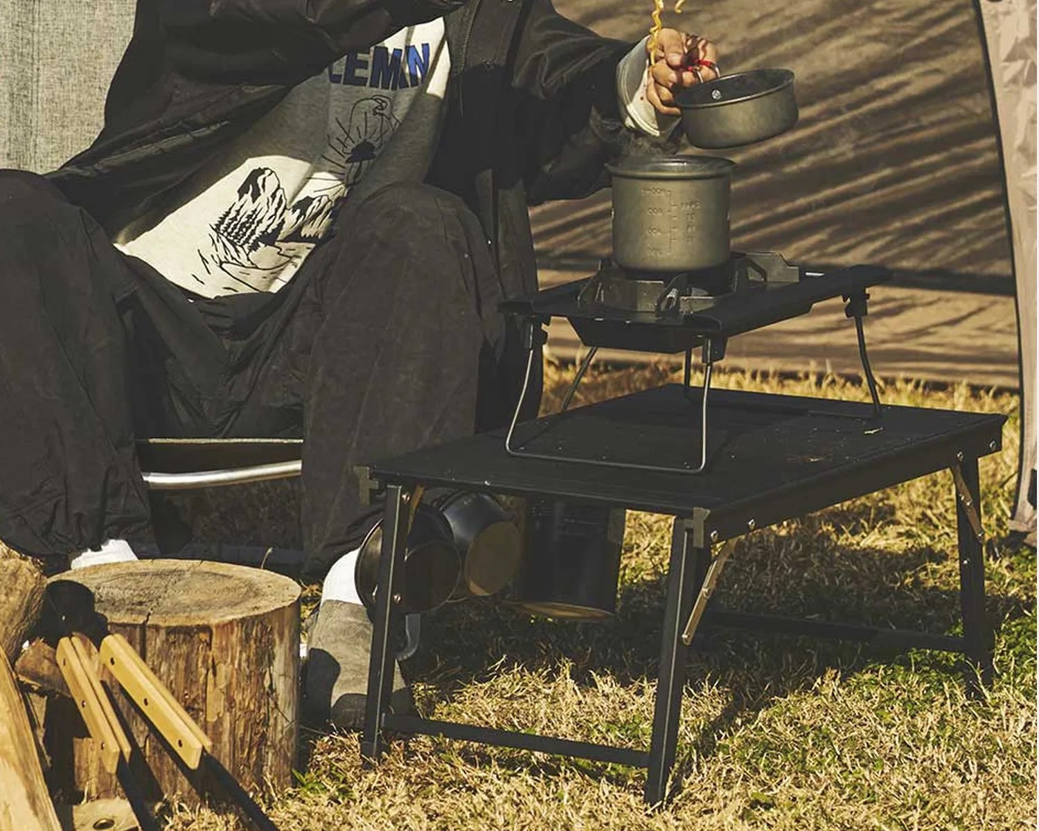 Black portable camping stove on a folding black metal table outdoors with a person lifting a pot lid.
