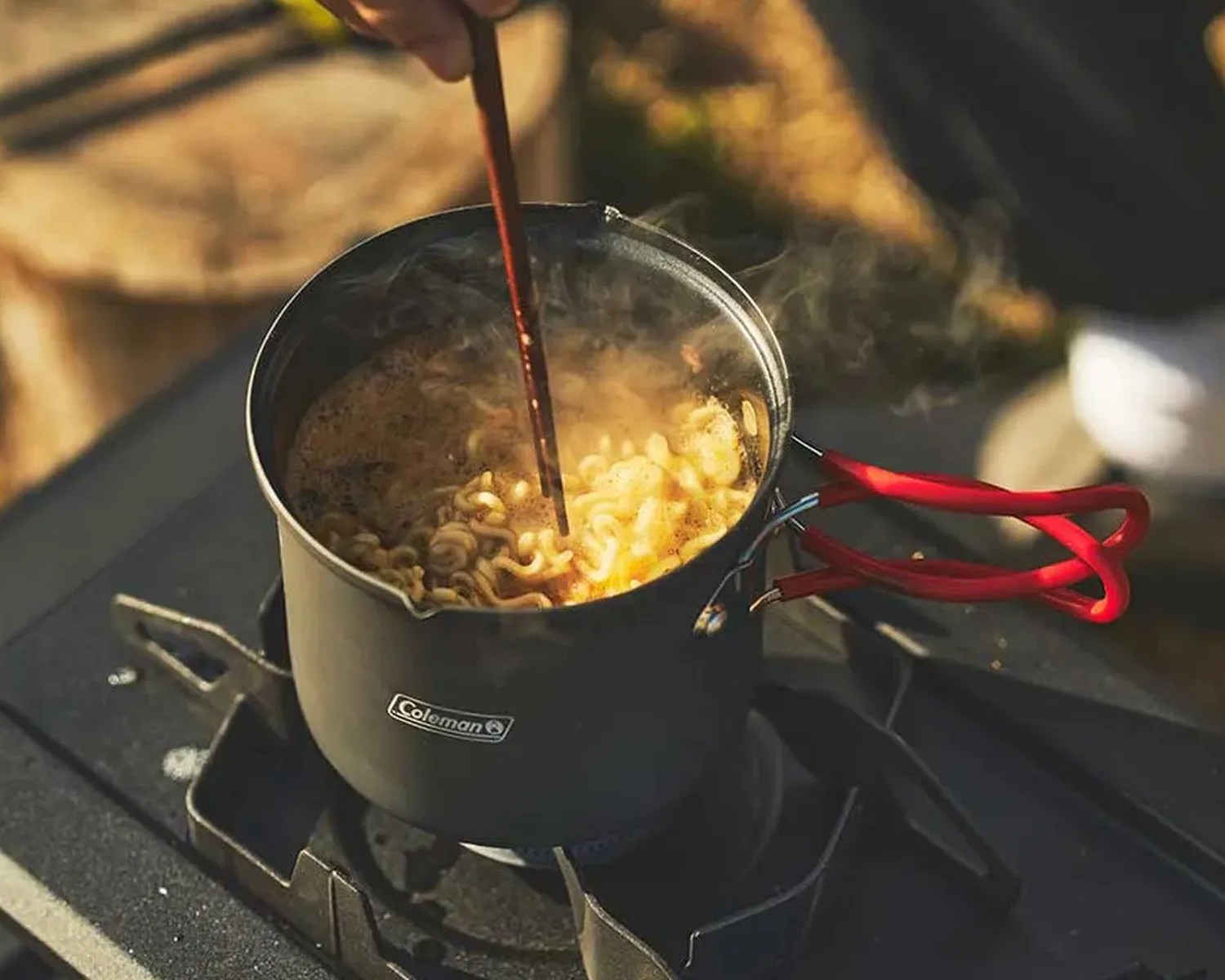 Black Coleman pot with red handles boiling instant noodles on a stove with steam rising.