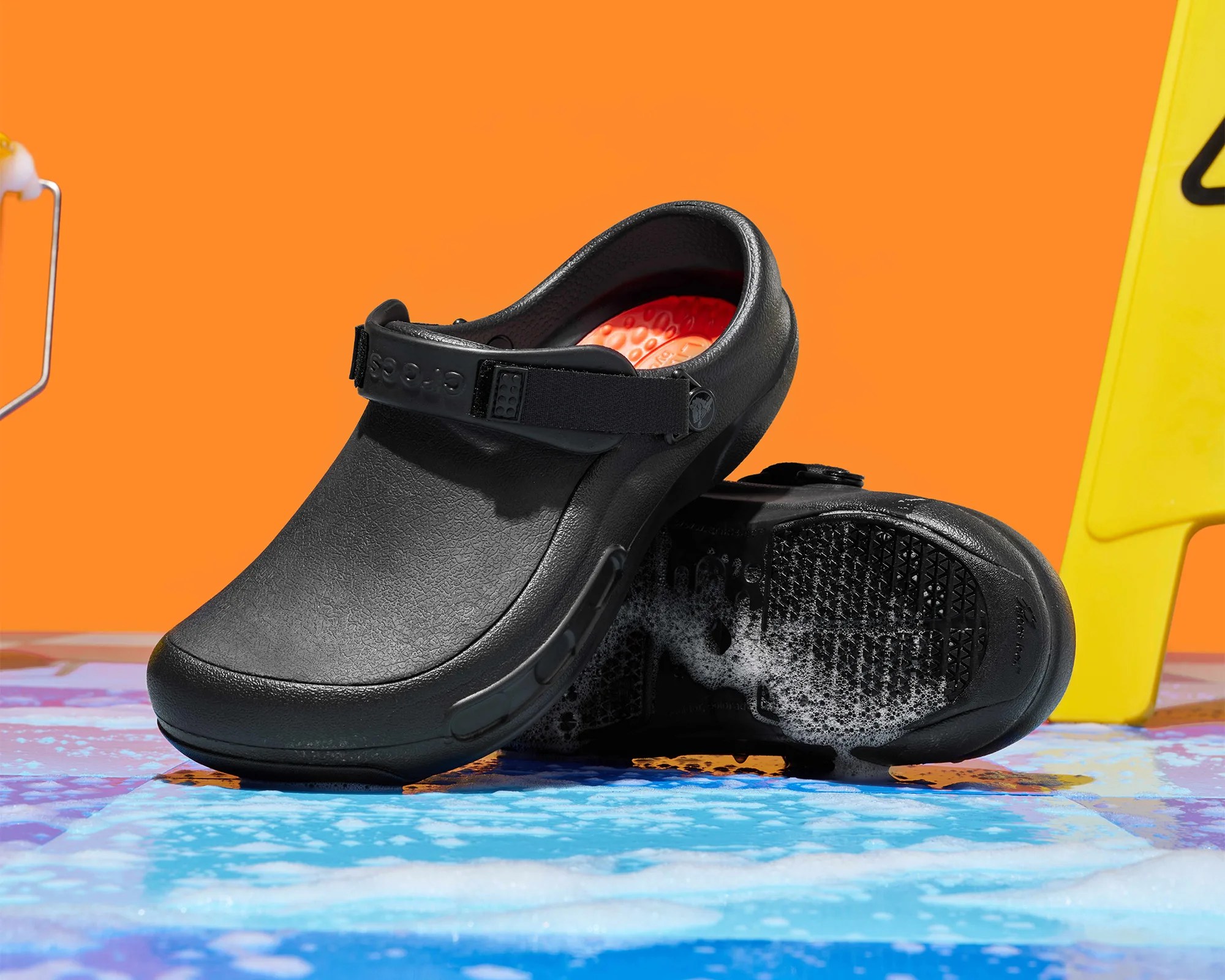 Black Crocs clog with an adjustable strap and orange textured insole on a wet tiled surface.