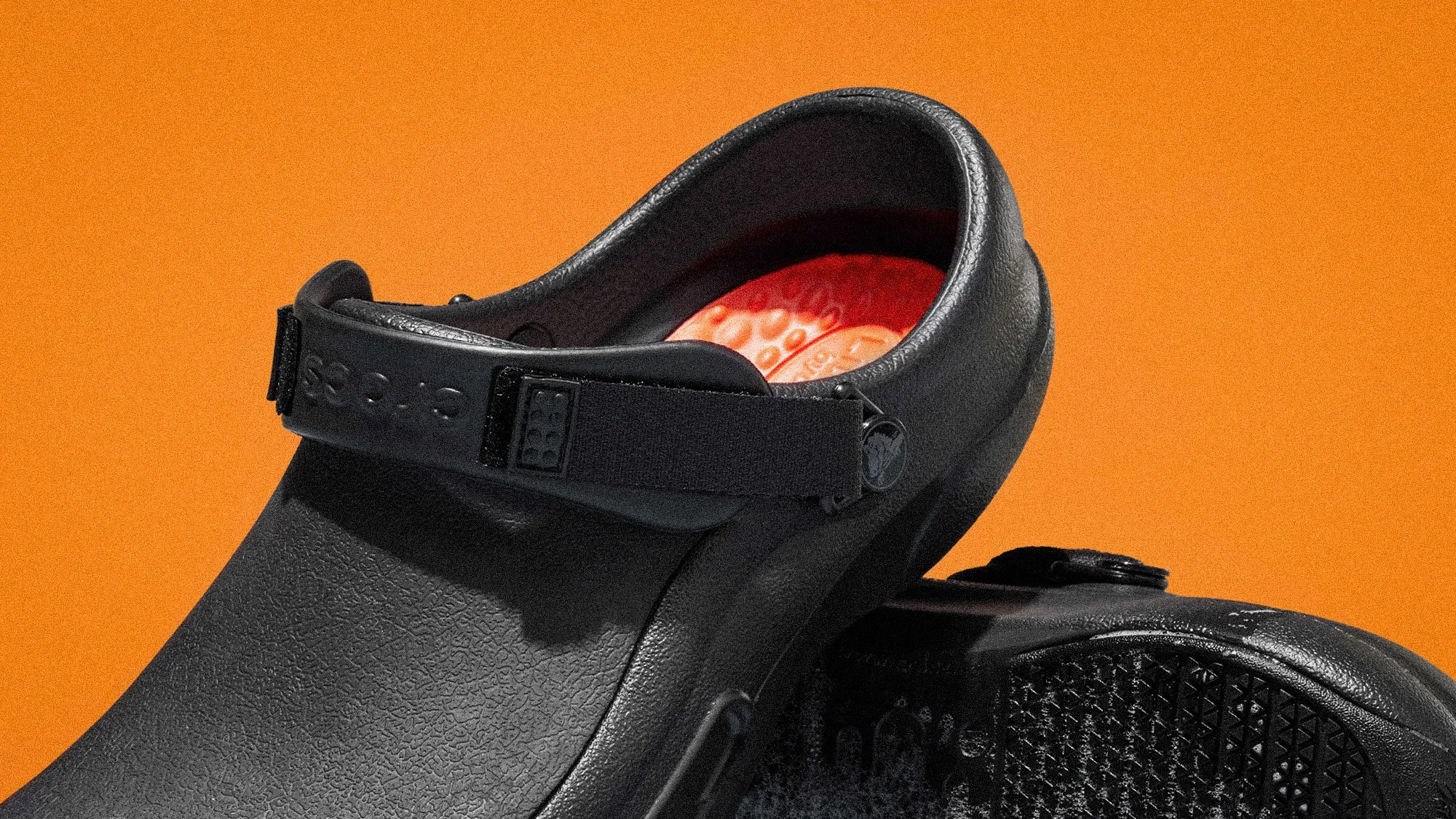 Black textured Crocs clog with adjustable heel strap and red textured insole against an orange background.