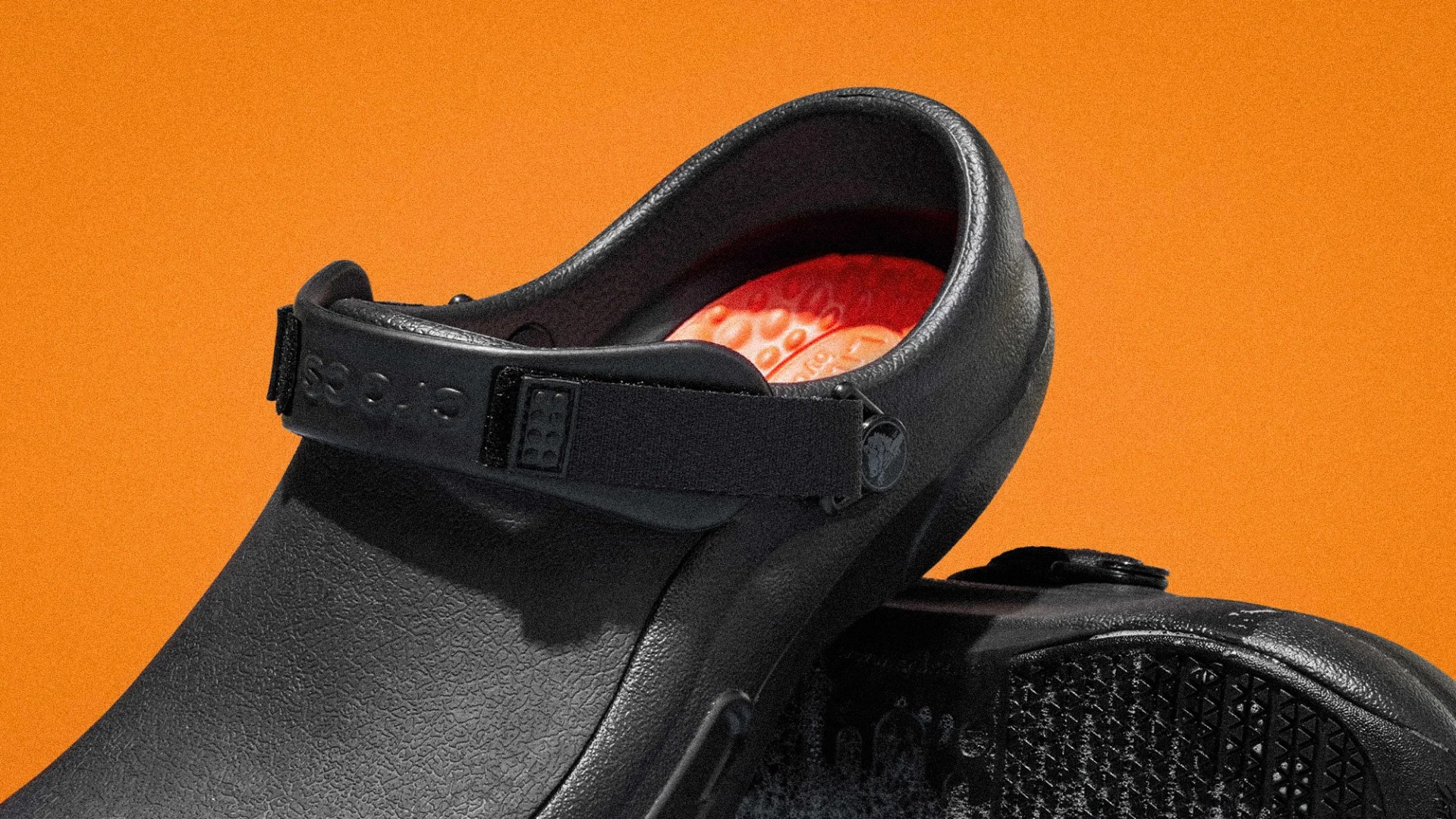 Black textured Crocs clog with adjustable heel strap and red textured insole against an orange background.