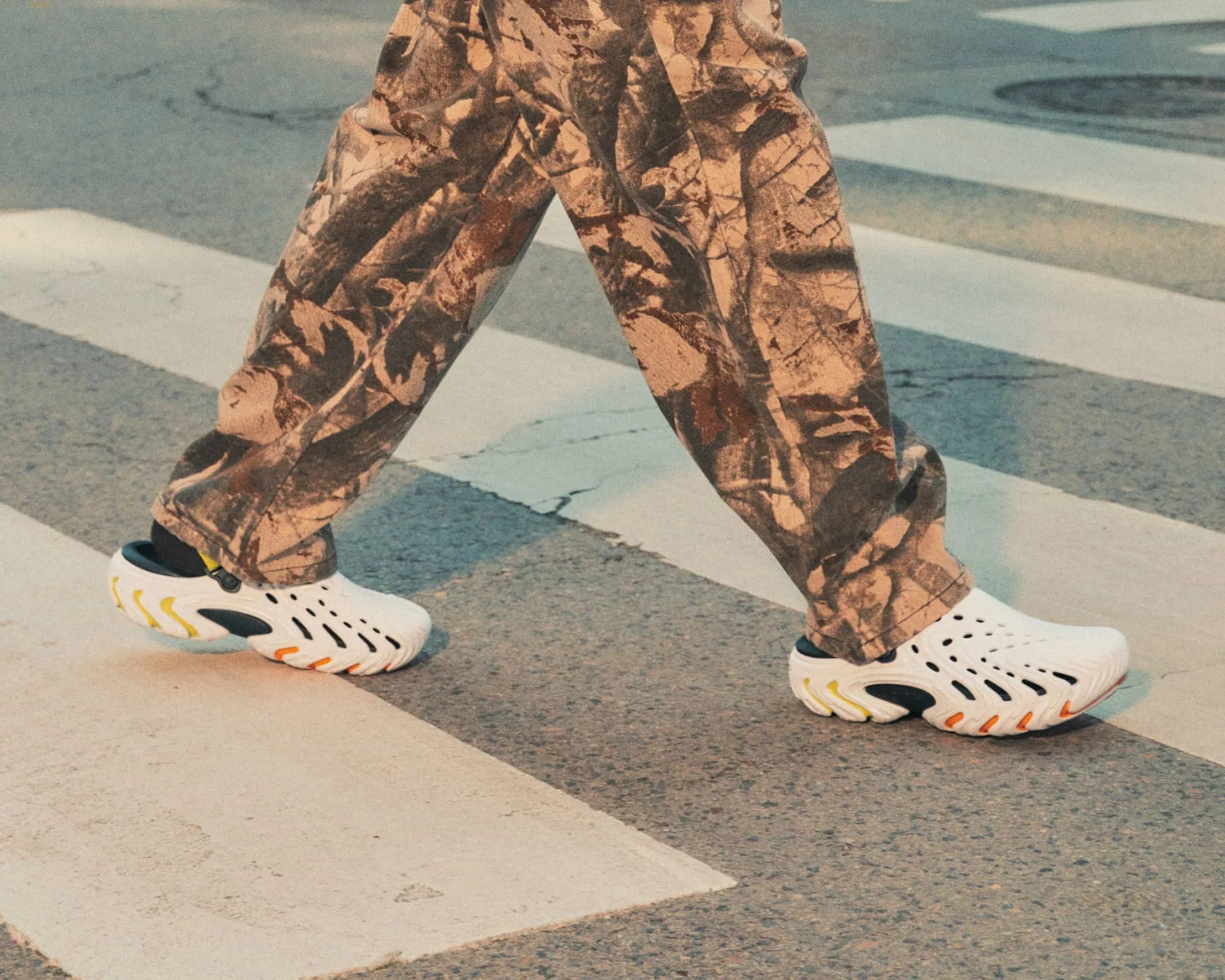 White slip-on shoes with black and yellow accents worn with brown camouflage pants on a street crosswalk.