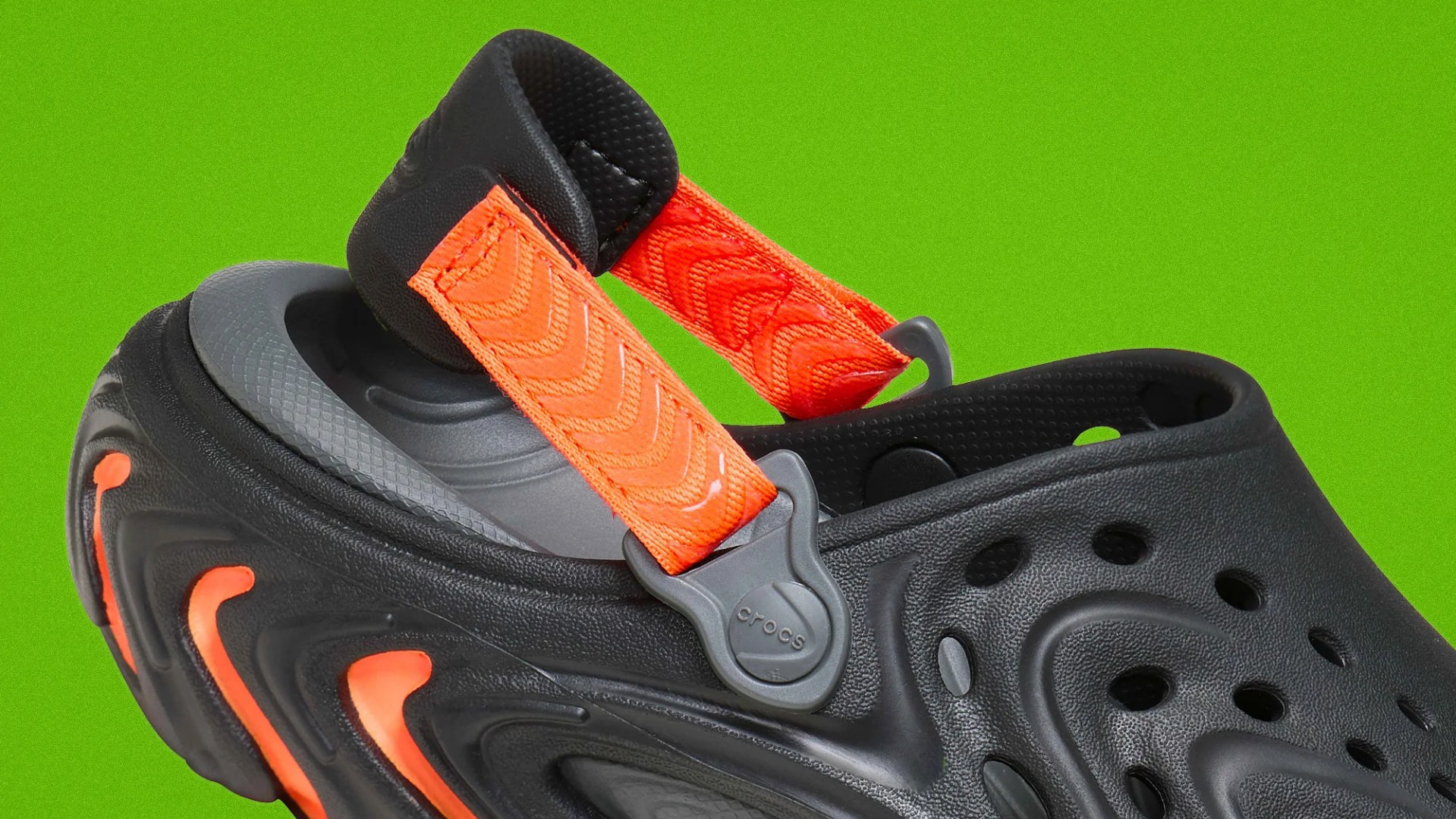 Black Crocs clog with orange accents and an adjustable orange strap on a green background.