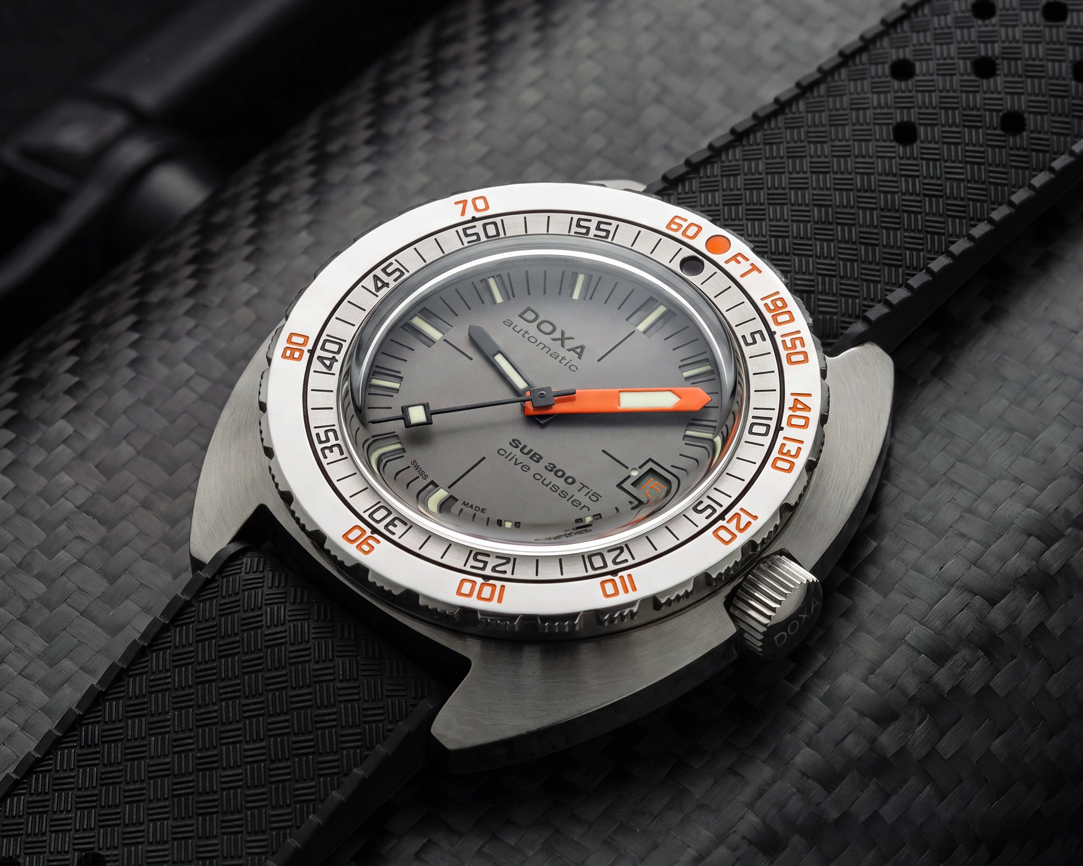 Close-up of a DOXA SUB 300 Ti5 dive watch with a silver bezel, orange minute hand, and black textured rubber strap on a carbon fiber surface.