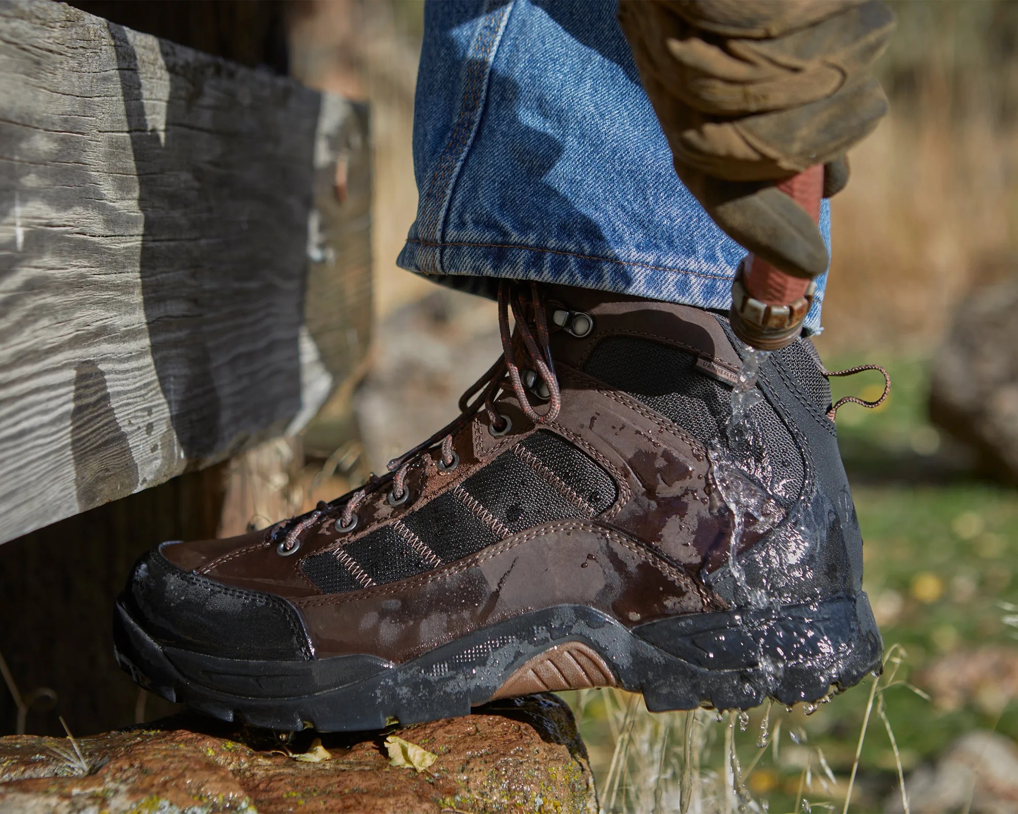 Brown and black waterproof hiking boot with water splashing on it, worn with blue jeans.