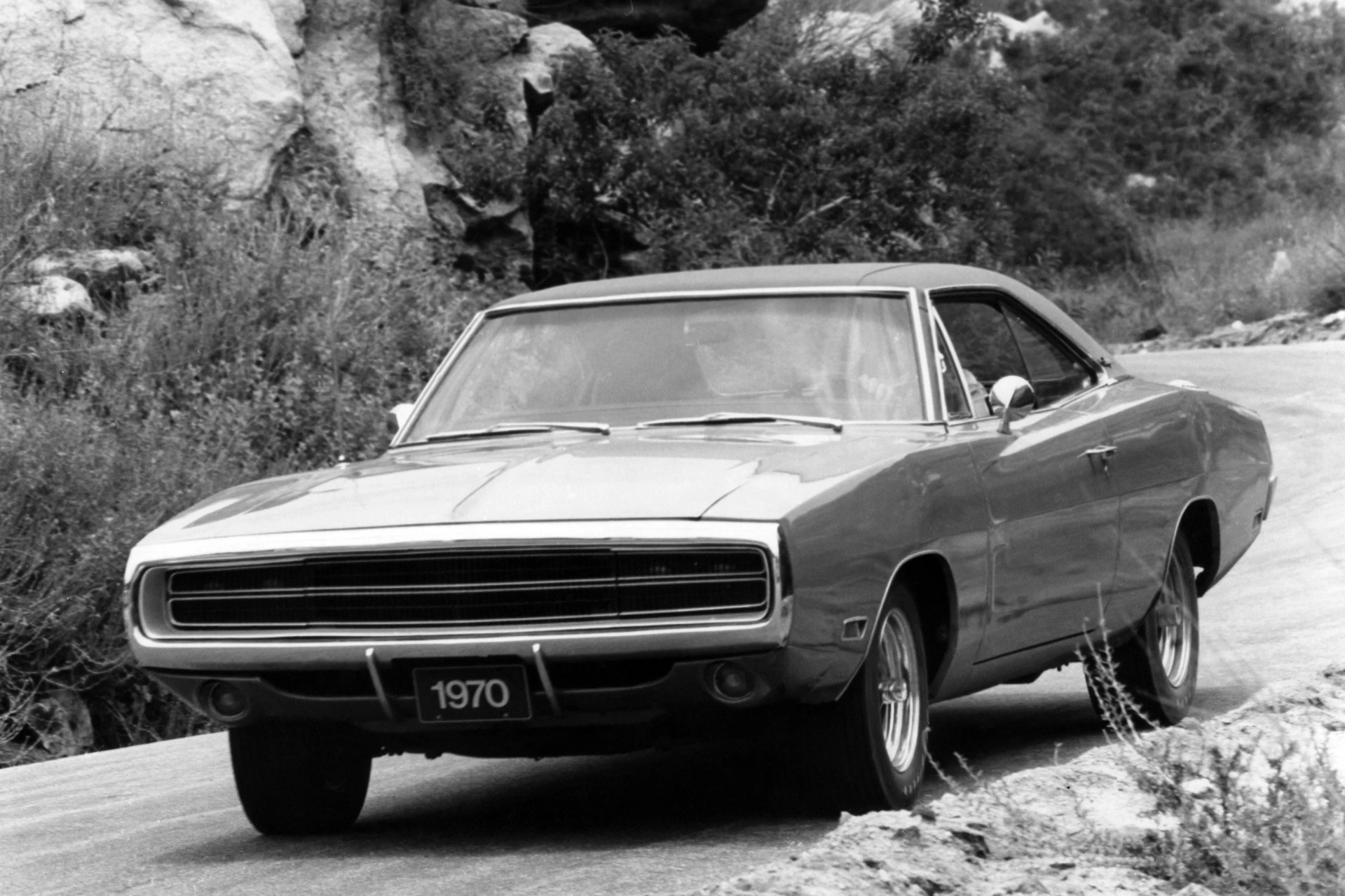 Black and white photo of a 1970 Dodge Charger driving on a winding road with rocky and bushy surroundings.