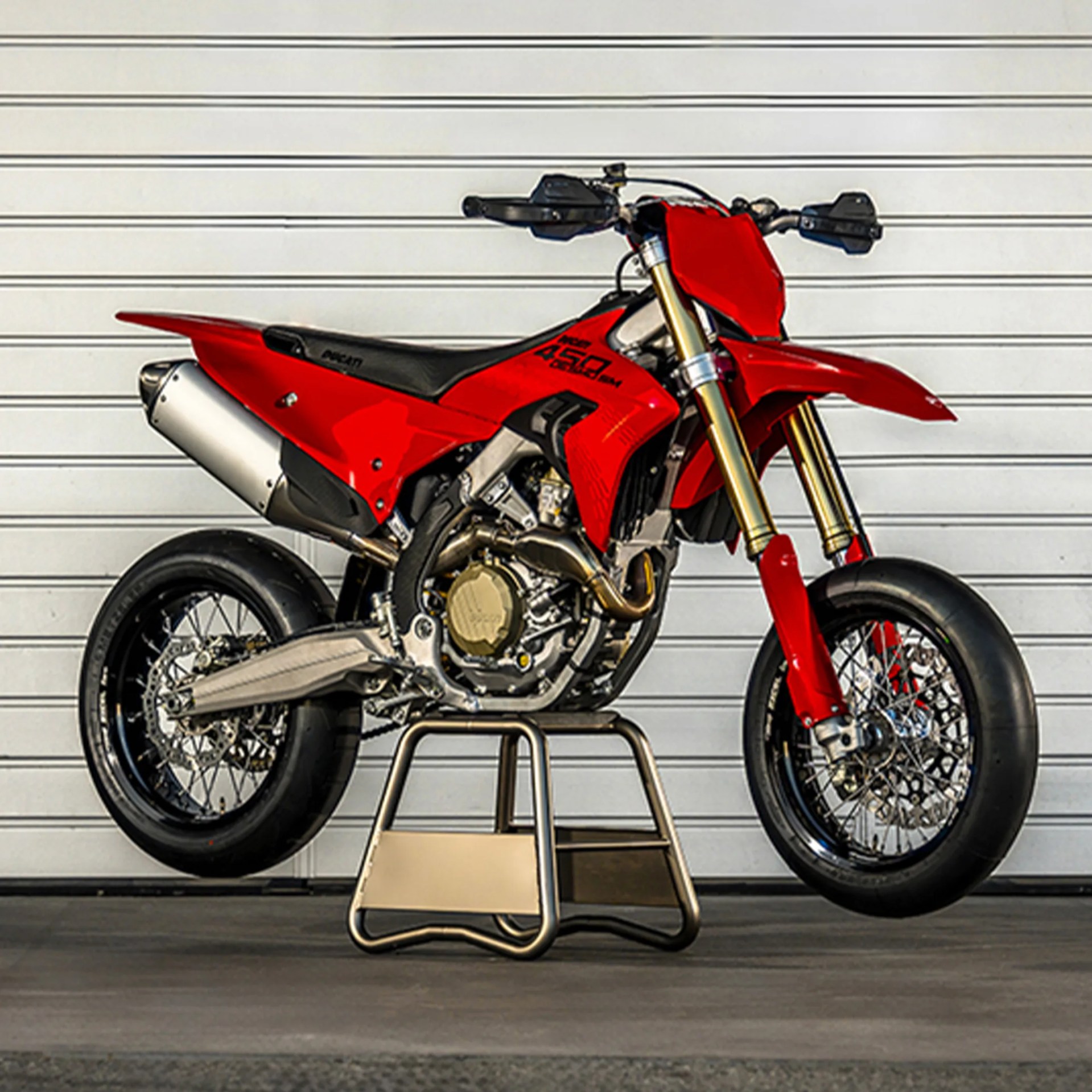 Red Ducati 450 SM motorcycle with gold front forks on a metal stand against a white garage door.