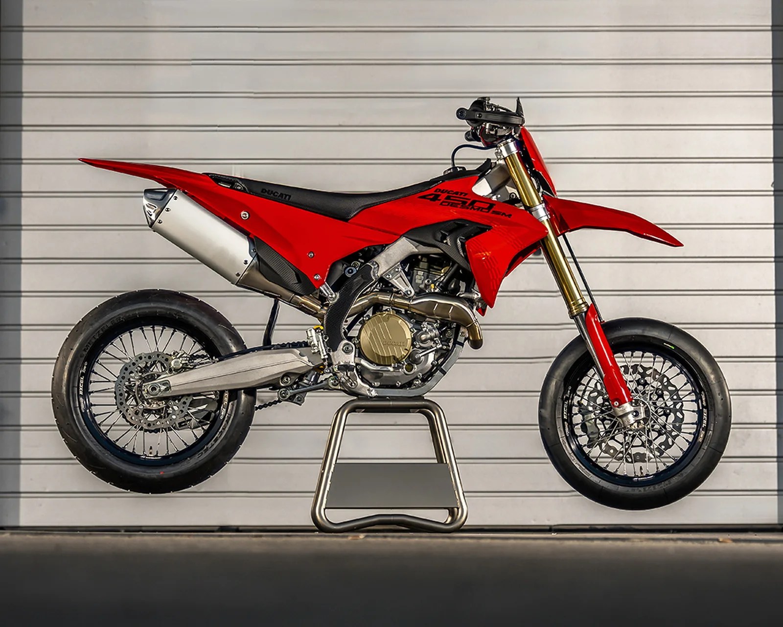 Red Ducati 450 SM motorcycle with black seat and gold front forks on a metal stand against a gray garage door.