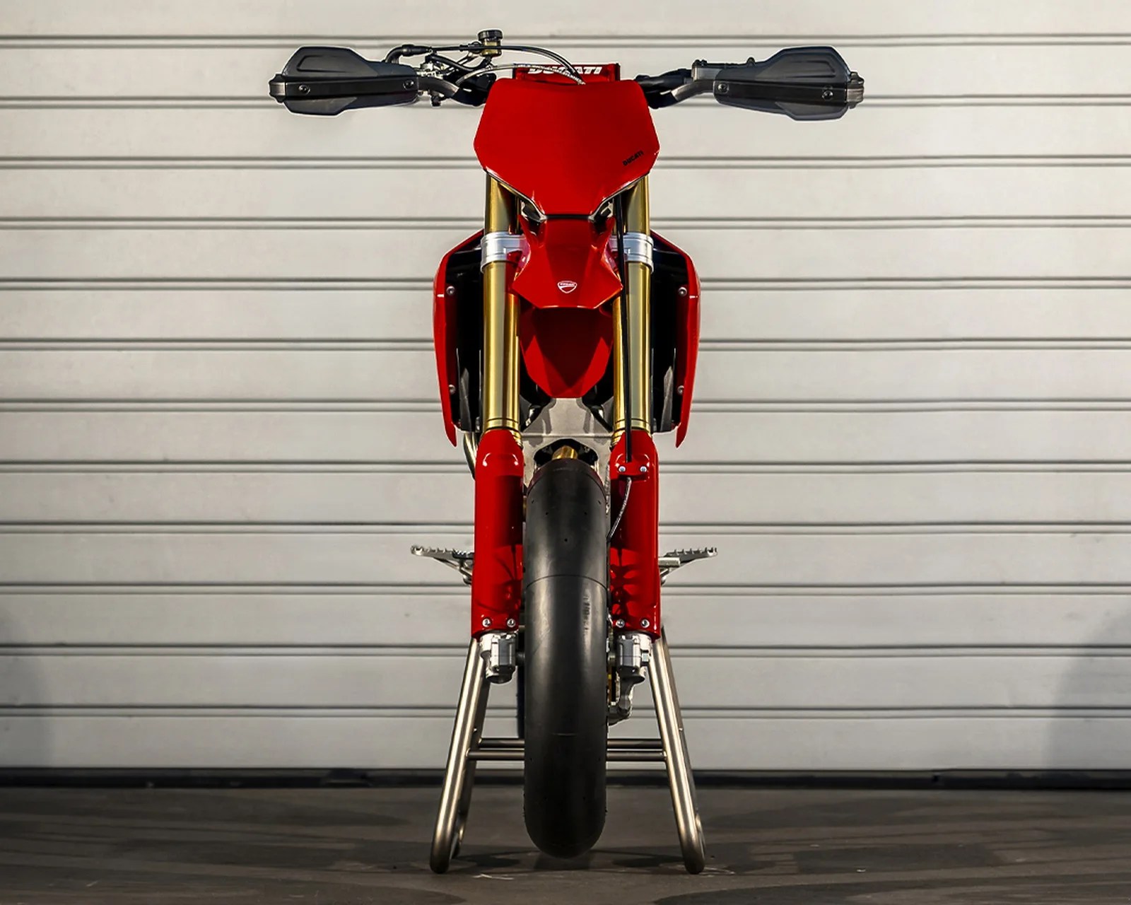 Front view of a red Ducati motorcycle with gold front forks and black handguards against a white garage door.