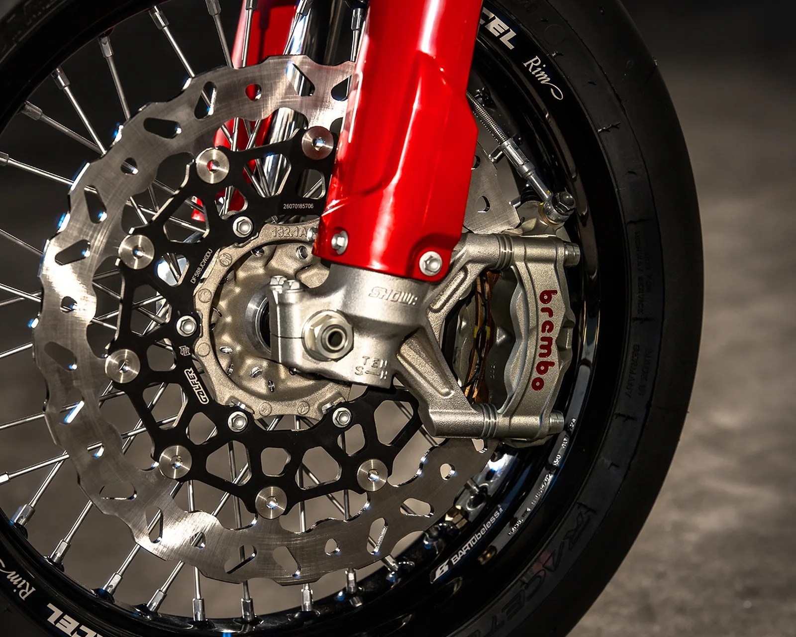 Close-up of a motorcycle front wheel showing a Brembo brake caliper, disc rotor, and red fork.
