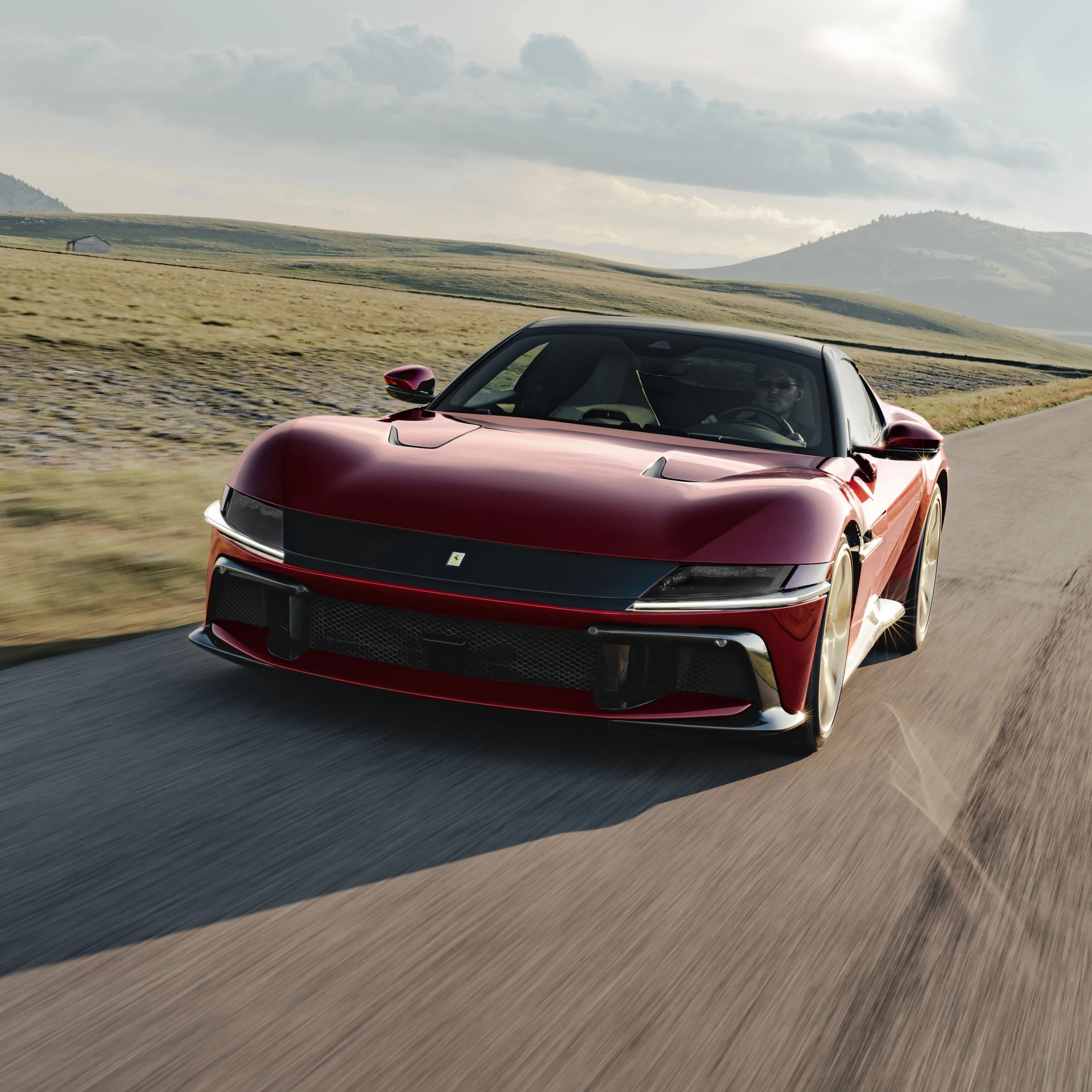 Red sports car driving on a rural road with grassy hills and cloudy sky in the background.