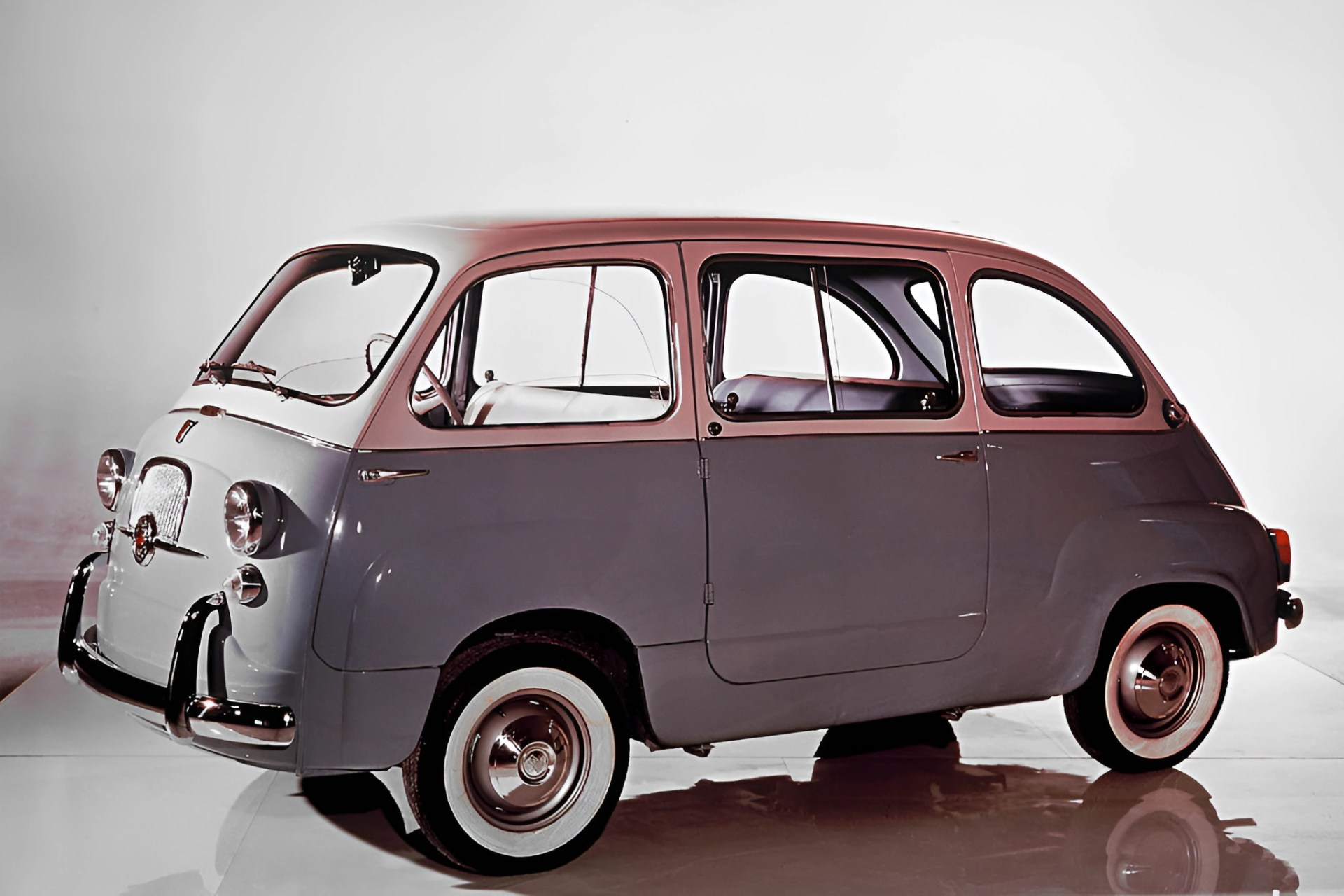Vintage compact gray car with rounded edges and whitewall tires on a reflective surface.
