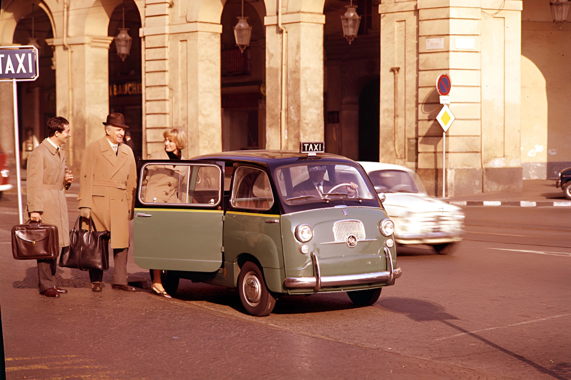 Green vintage taxi with black roof and open door parked on street near beige stone building with three people in coats carrying bags.