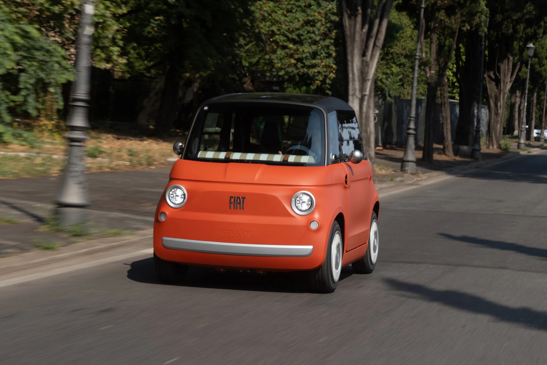 Compact matte orange Fiat Topolino car with black roof driving on a tree-lined street.
