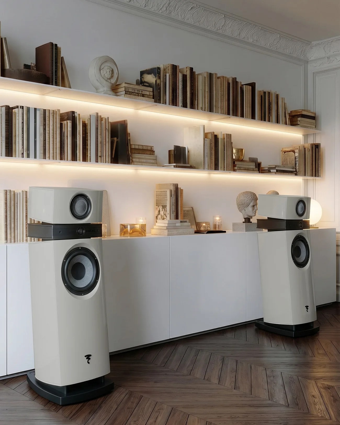 Two large white floor-standing speakers with black bases in a modern room with white shelves filled with books and decorative busts.