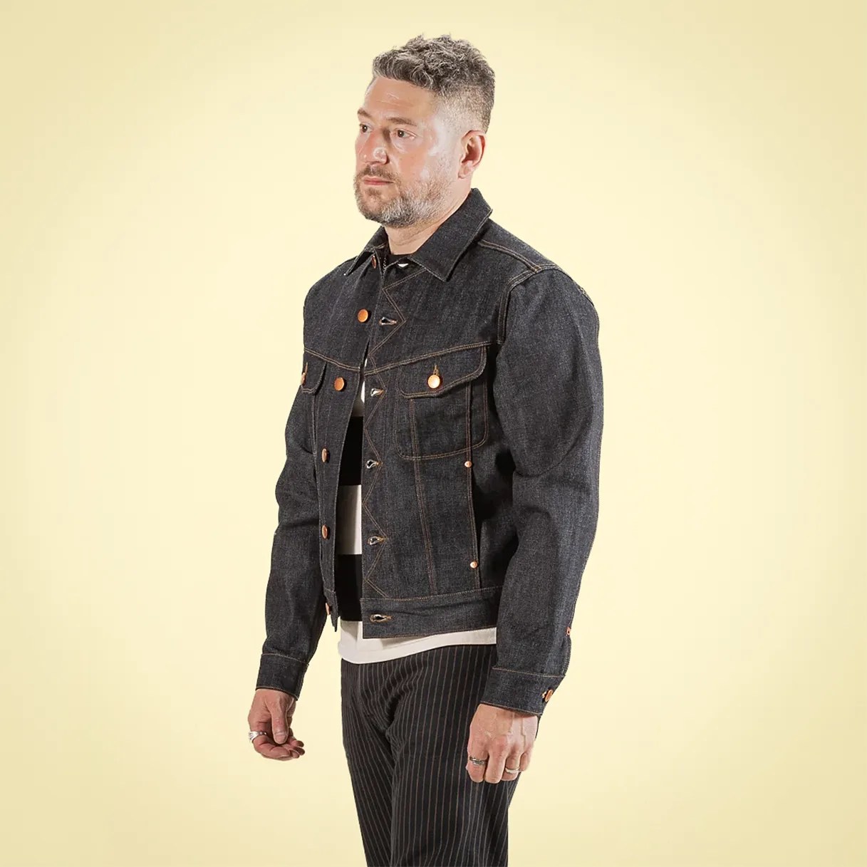 Man wearing a dark denim jacket with copper buttons and contrast stitching against a light yellow background.