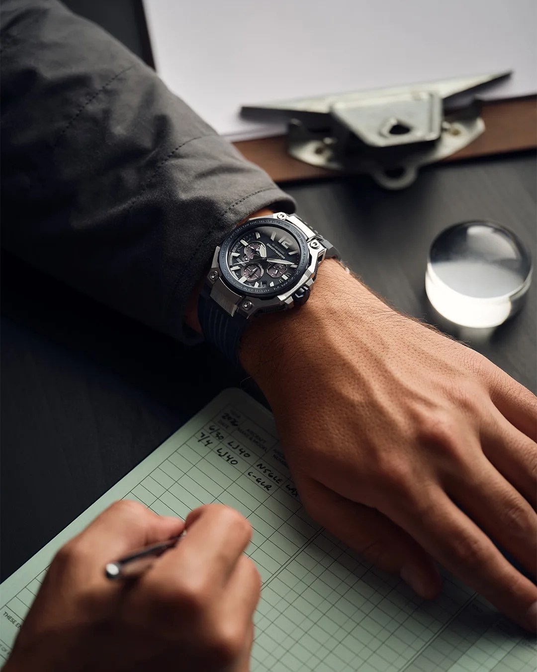 Black and silver chronograph watch with a black rubber strap worn on a wrist over a dark jacket sleeve.