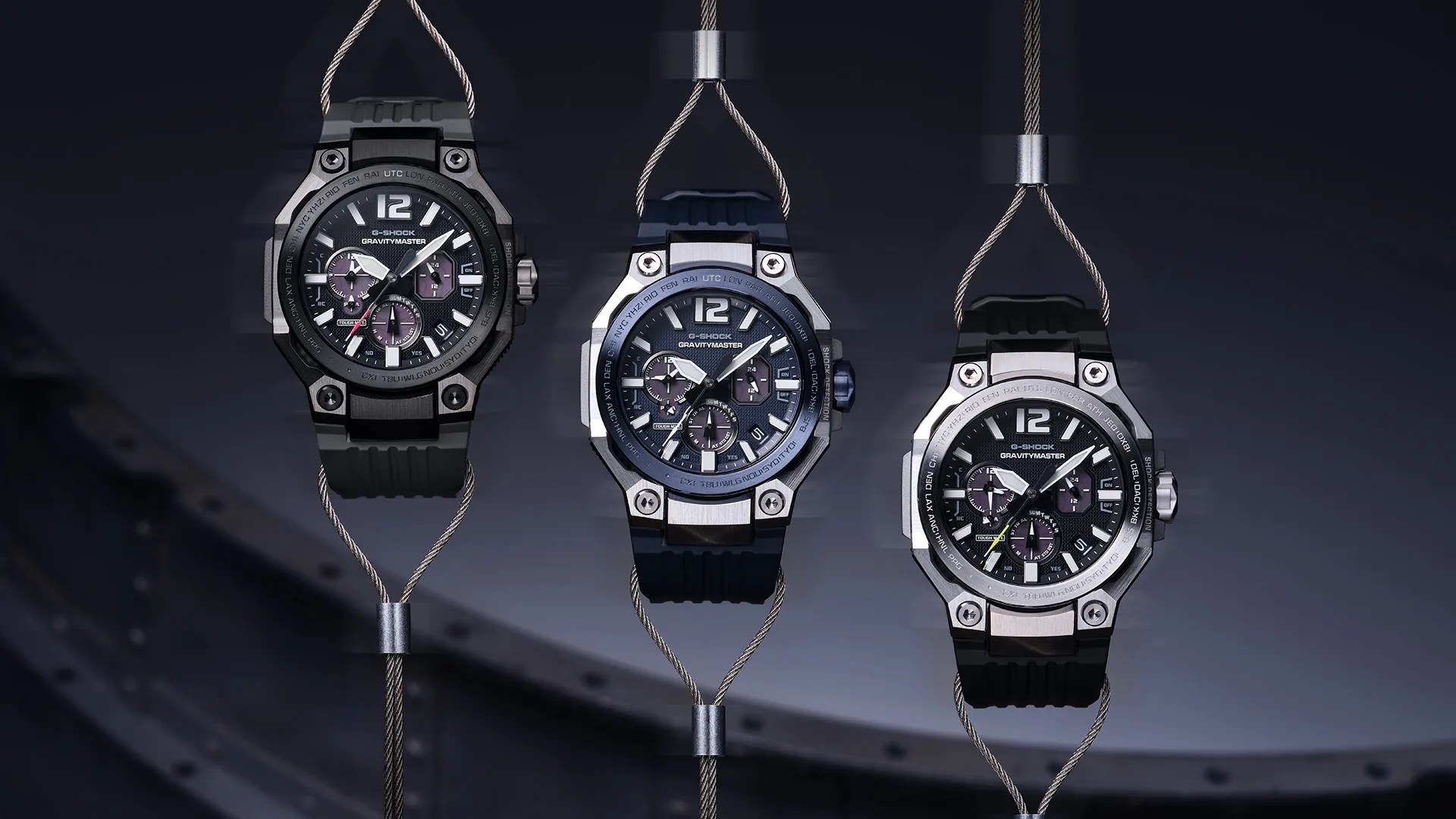 Three G-Shock Gravitymaster watches with black bands and multi-function dials hanging on metal cables against a dark background.
