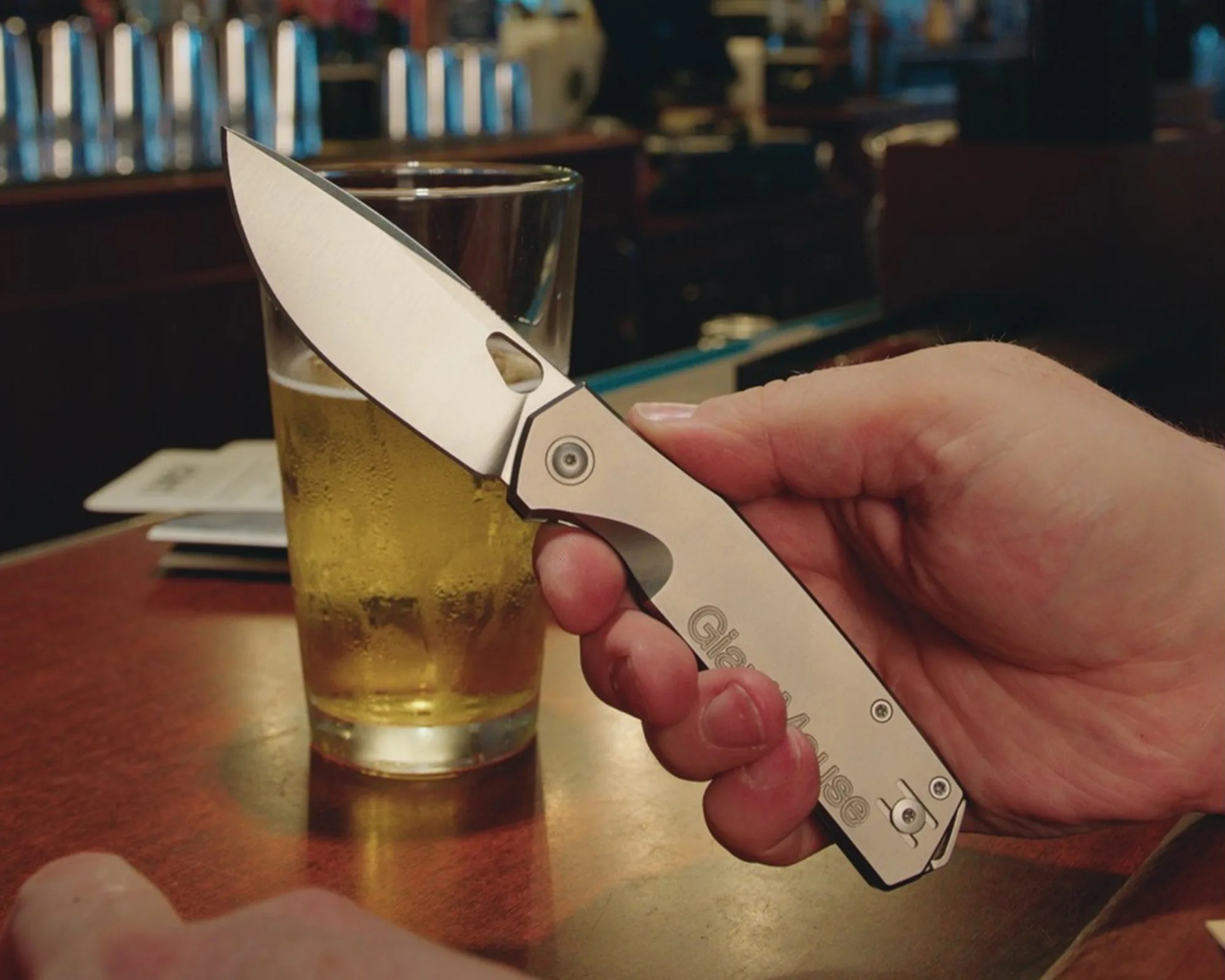 Hand holding a silver folding knife with a partially serrated blade over a glass of beer on a wooden table.