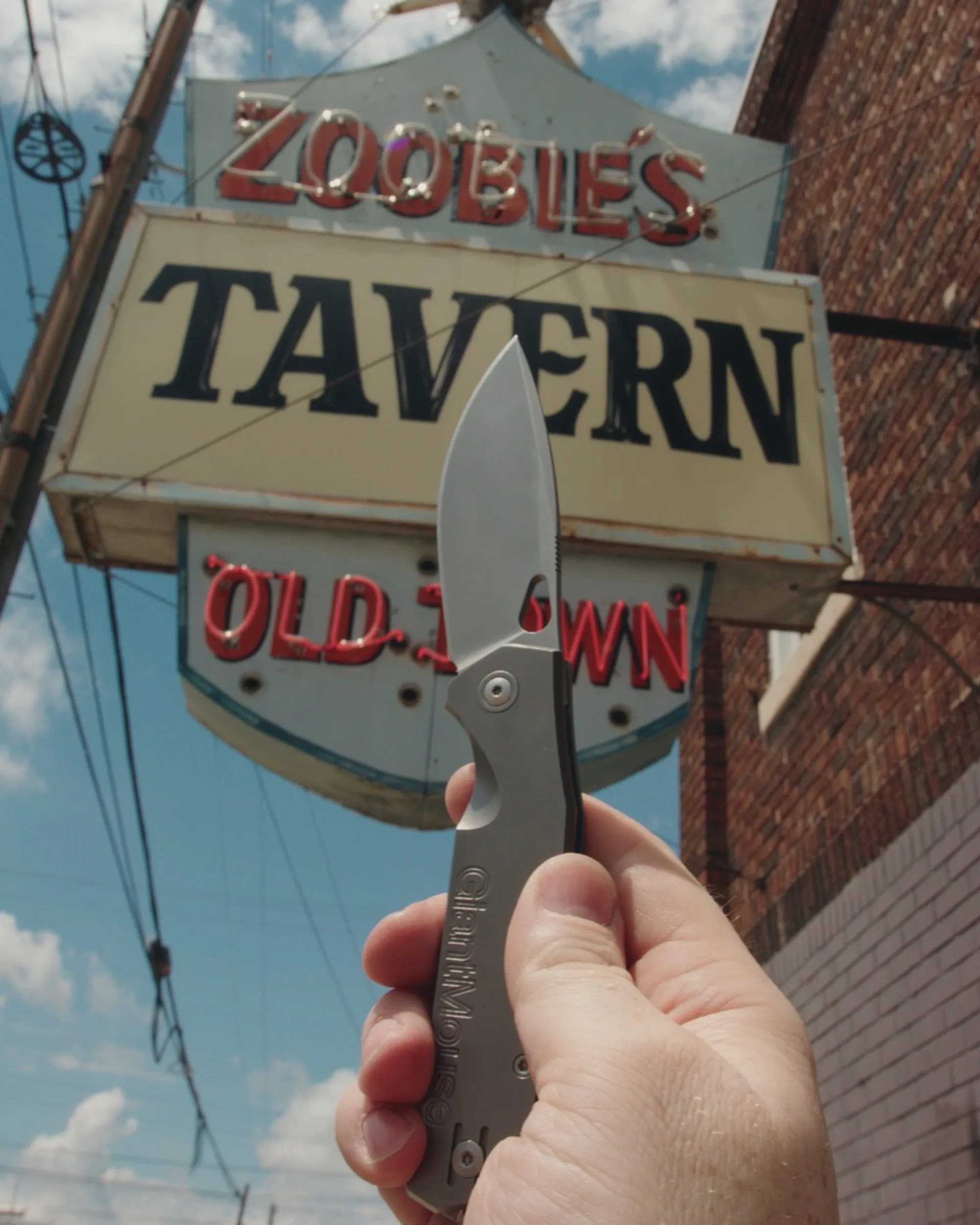 Hand holding a gray folding knife with a partially serrated blade in front of a Zoobilee's Tavern sign.