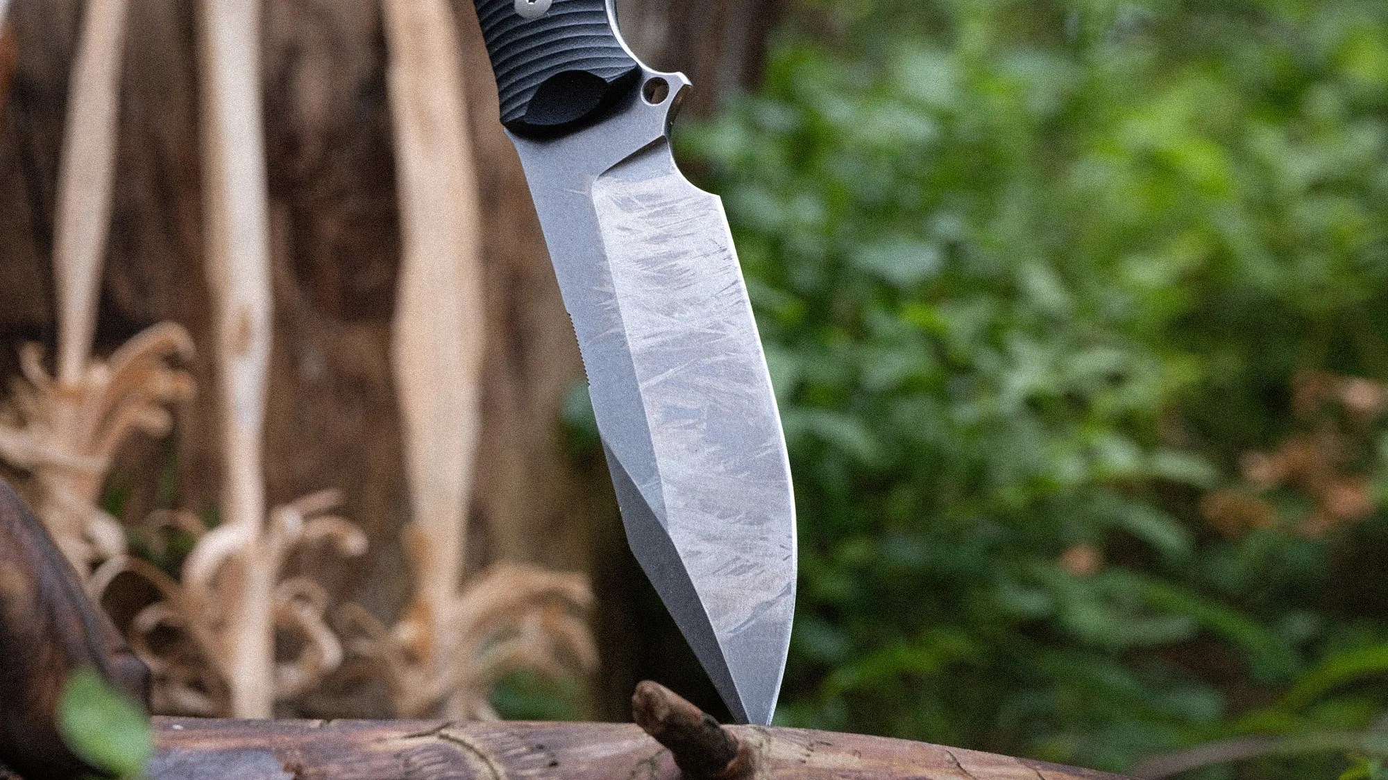 Knife with a textured black handle and a scratched, stonewashed blade stuck into a piece of wood outdoors.