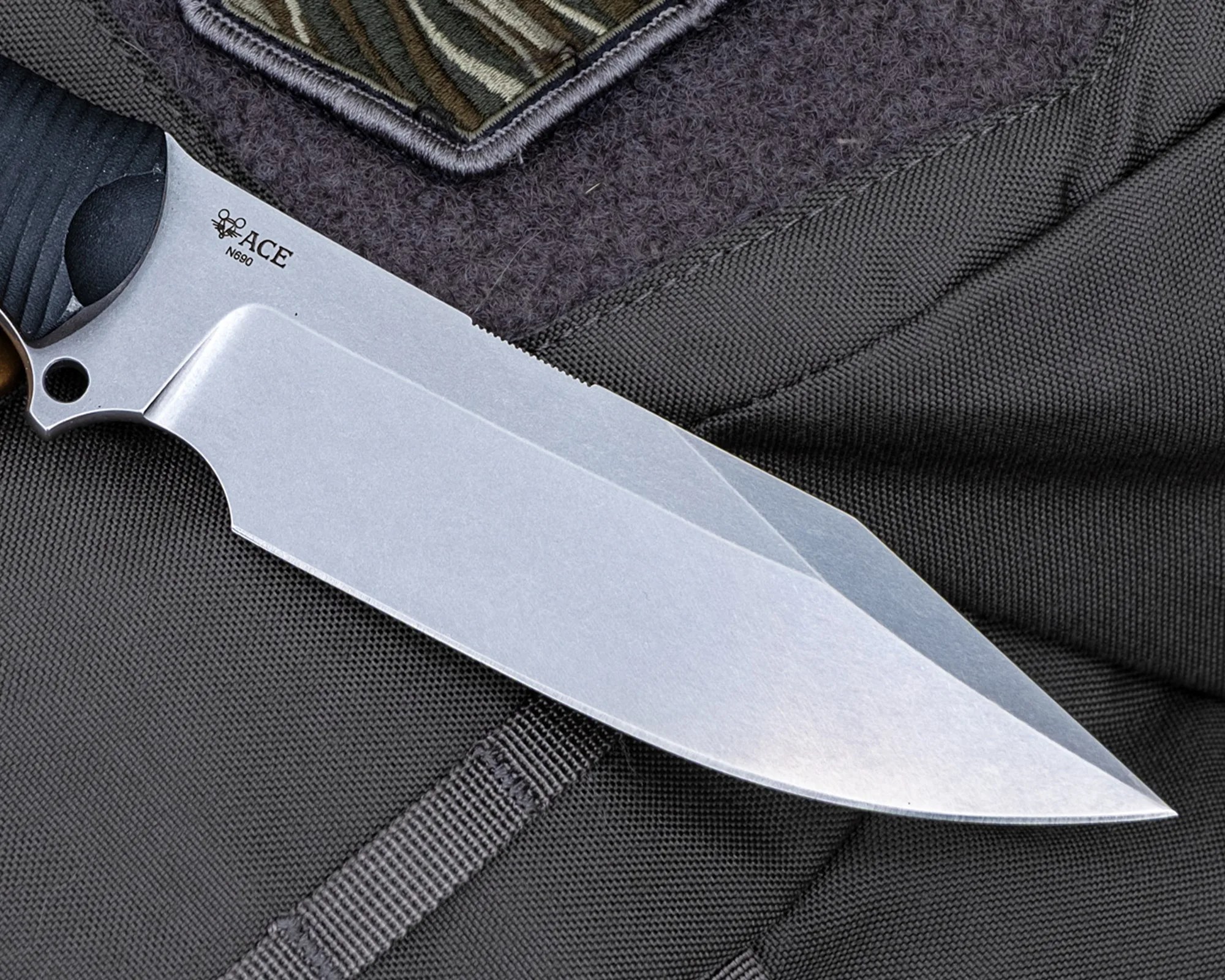 Close-up of a silver ACE N690 knife blade with a black textured handle on dark fabric.