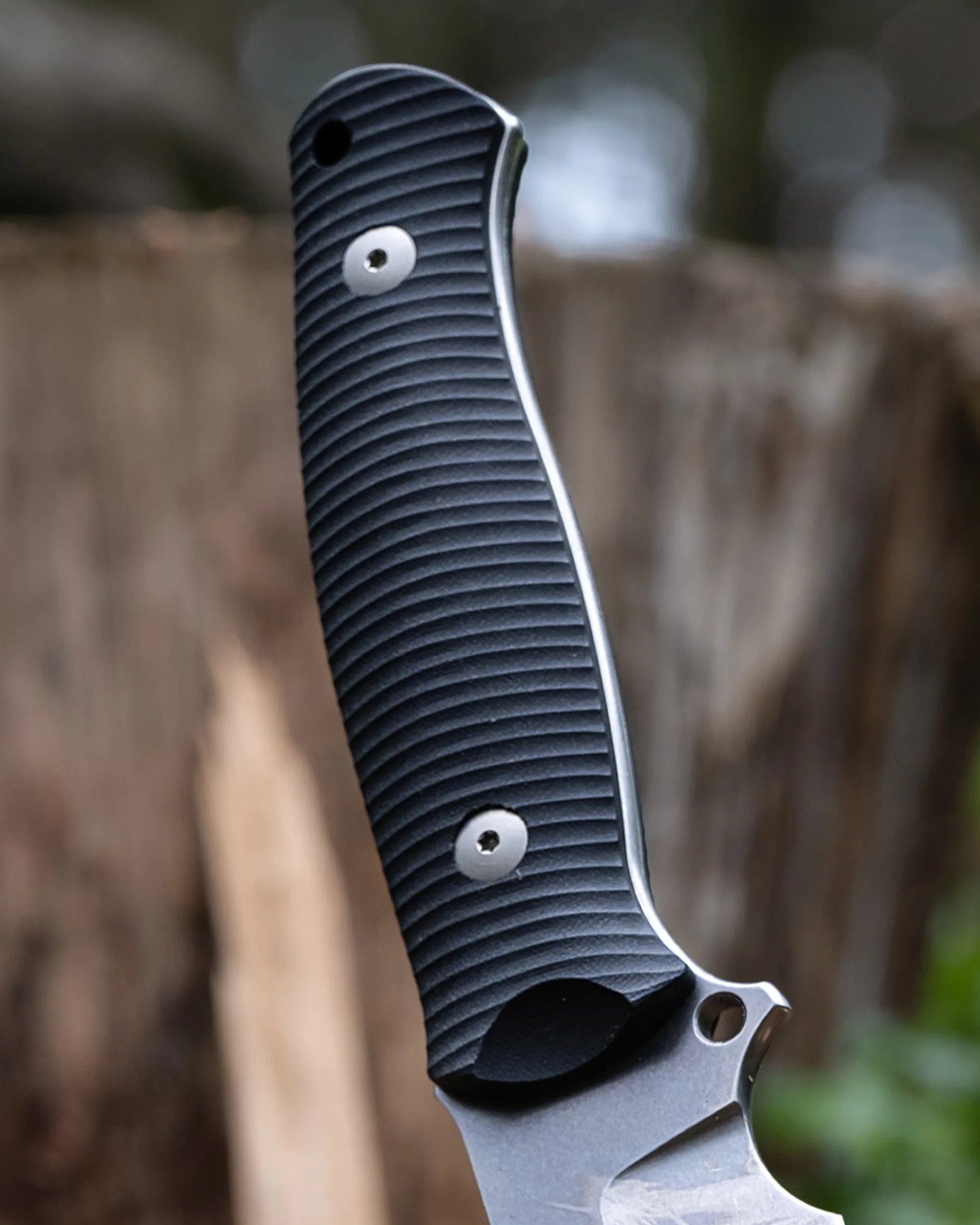 Black textured knife handle with silver screws and a partially visible blade against a blurred natural background.