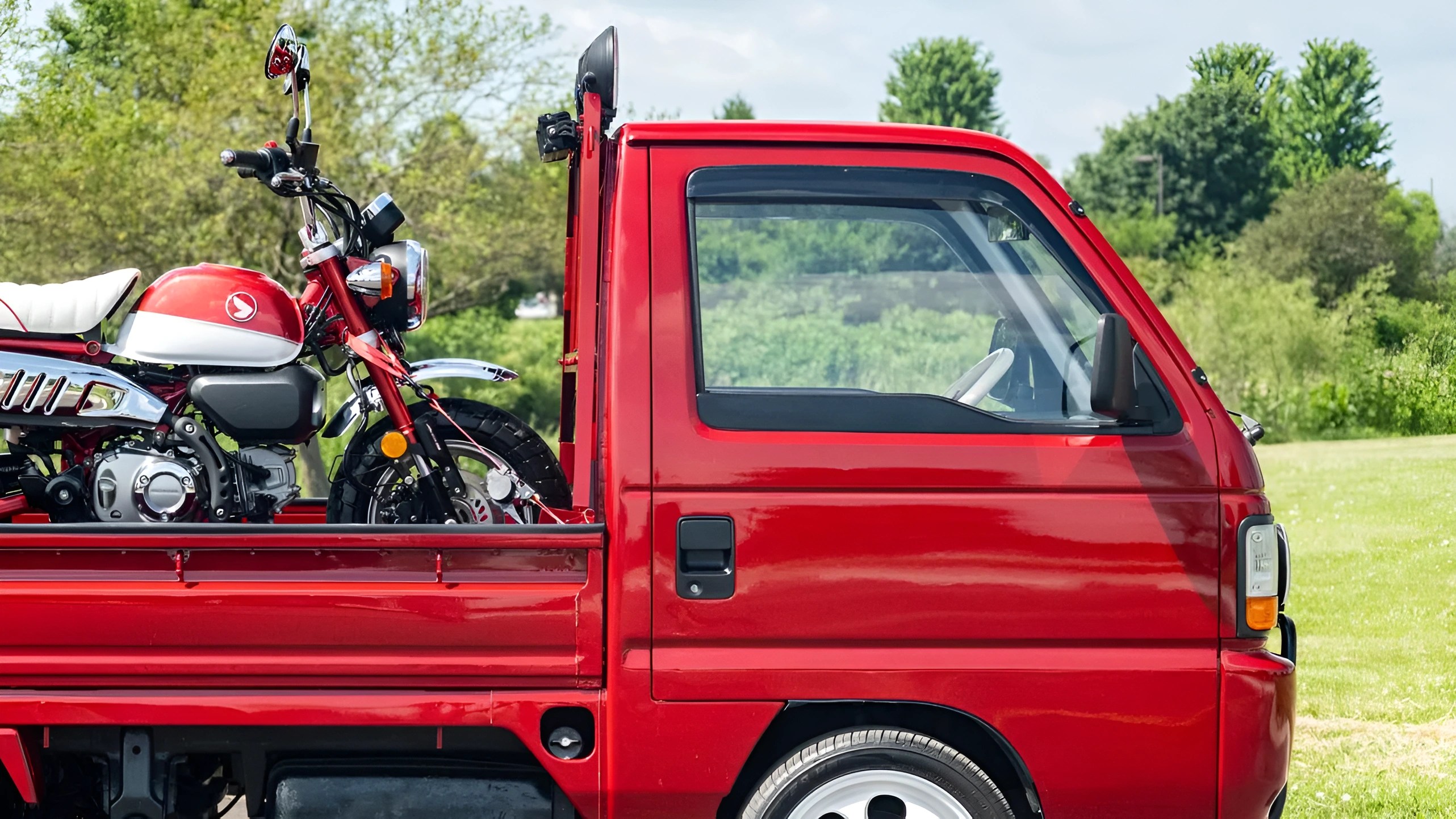 Red mini truck with a red and white motorcycle loaded in the truck bed, parked outdoors with greenery in the background.
