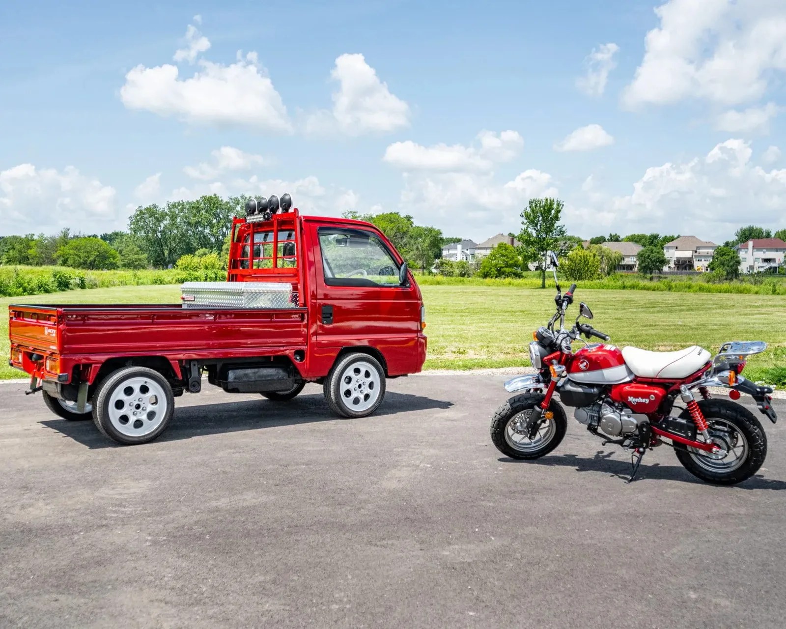 Red mini truck with silver toolbox in bed parked on pavement next to red and white Honda Monkey motorcycle.