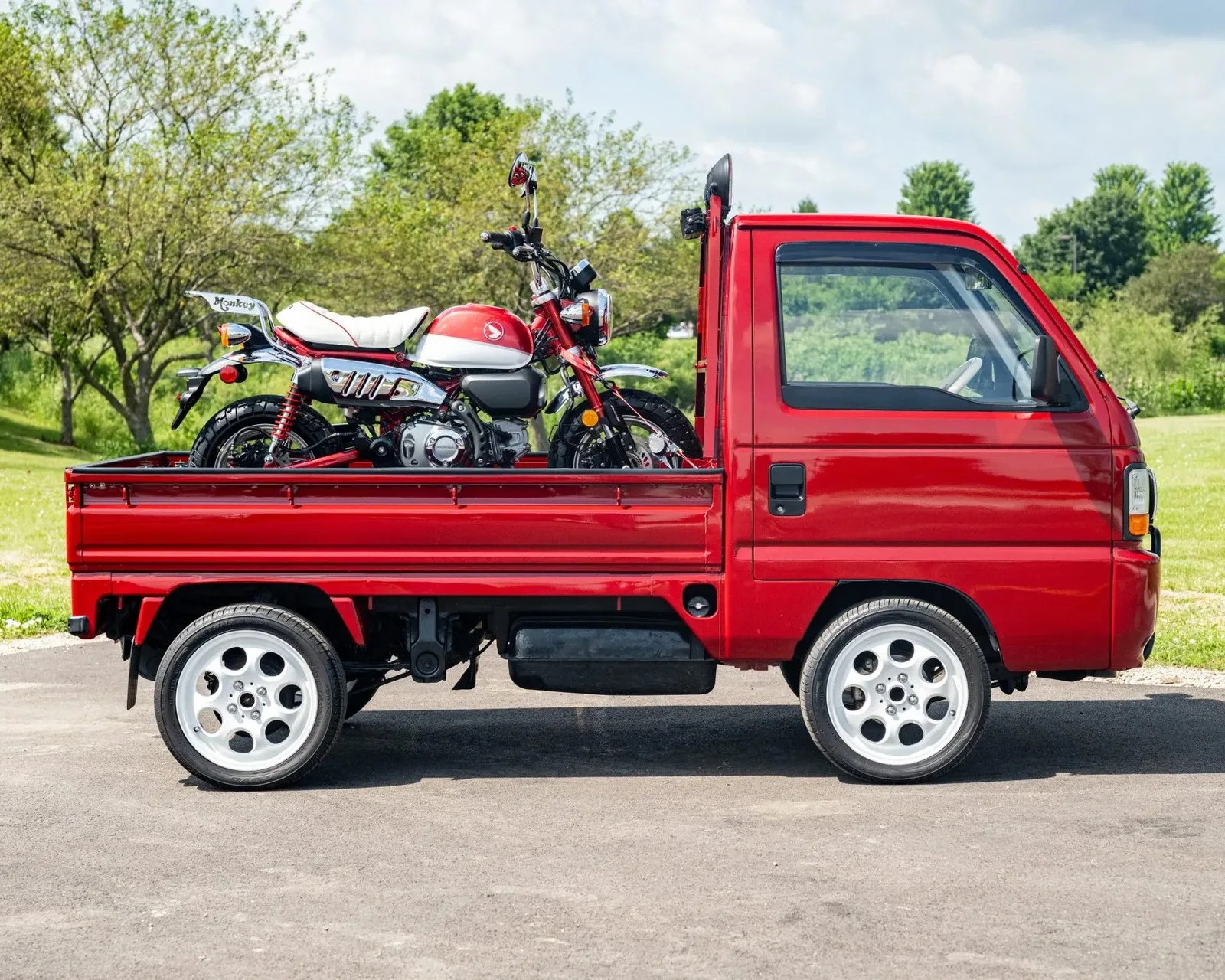 Red mini truck with white wheels carrying a red and white Honda Monkey motorcycle in its bed.