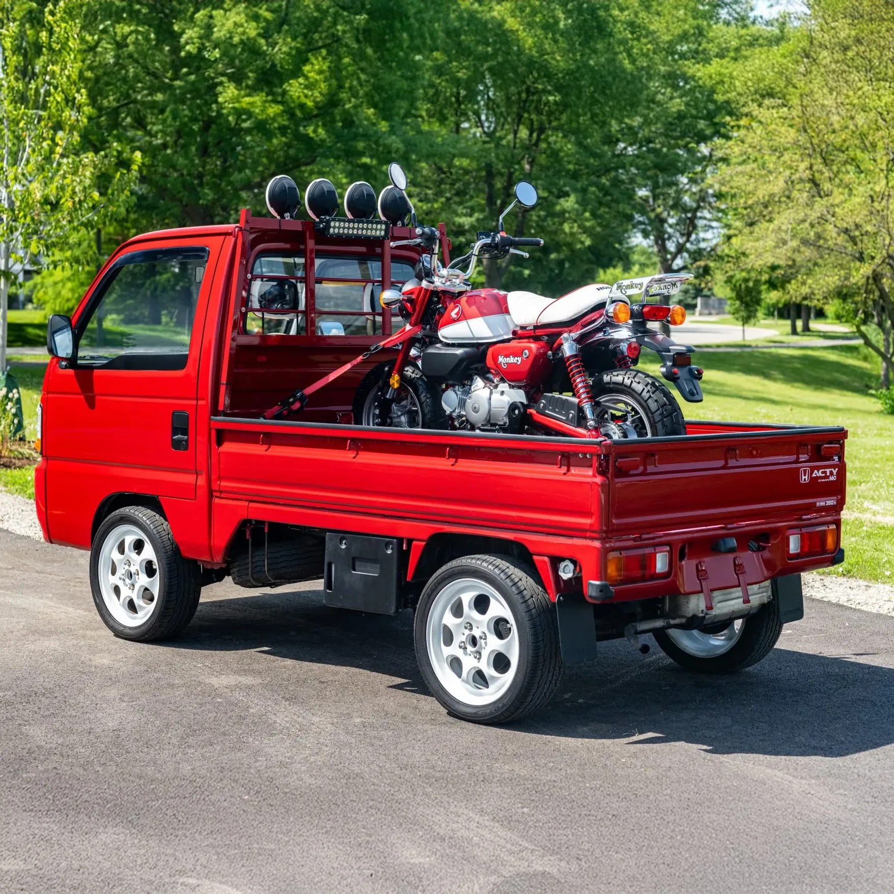 Red Honda Acty mini truck with a red and white Honda Monkey motorcycle secured in the truck bed, parked outdoors near green trees.