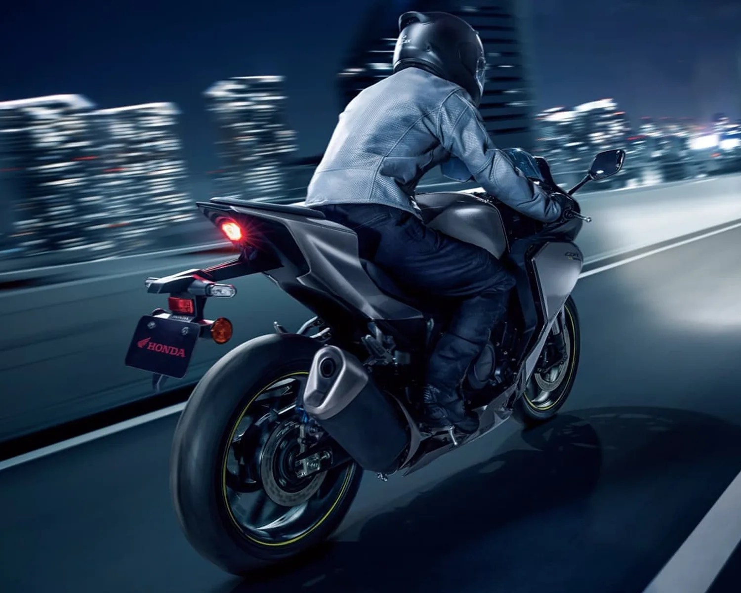 Motorcyclist in black helmet and jacket riding a silver Honda sportbike on a city road at night.
