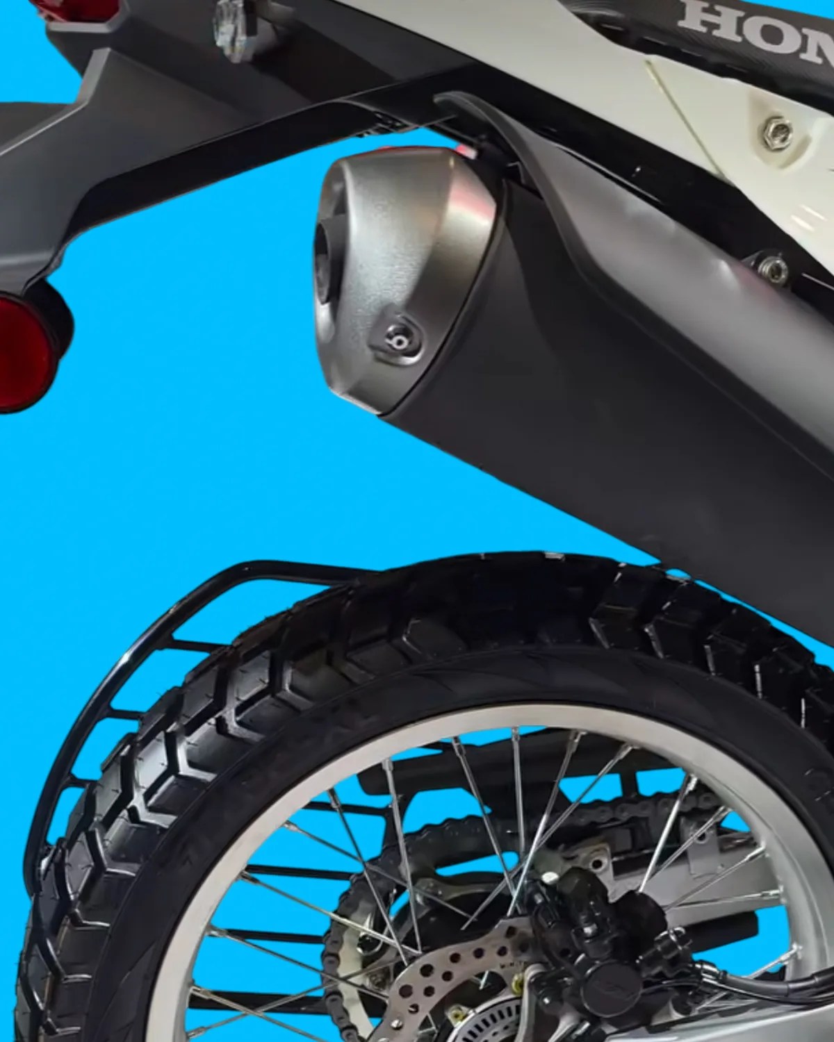 Rear section of a Honda motorcycle showing the exhaust pipe, rear tire with knobby tread, and part of the brake system.