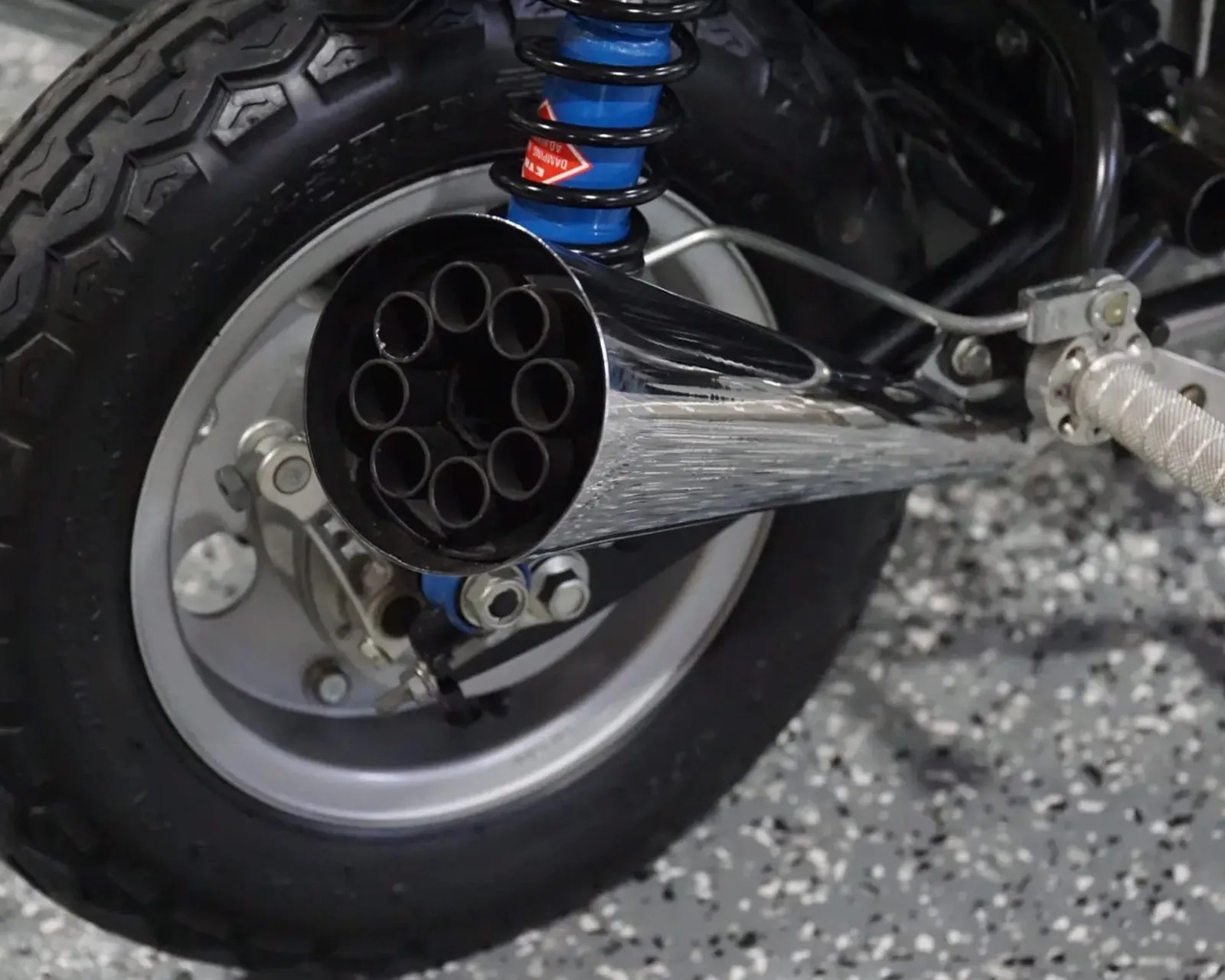 Close-up of a motorcycle rear wheel with a silver multi-pipe exhaust and blue coil spring suspension.