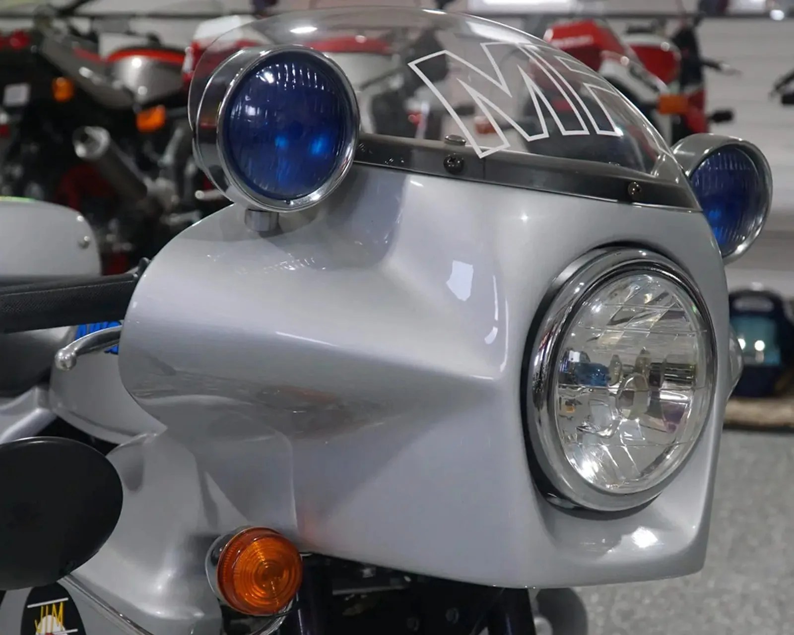 Close-up of a silver motorcycle front fairing with a clear headlight, two blue auxiliary lights, and an orange turn signal.