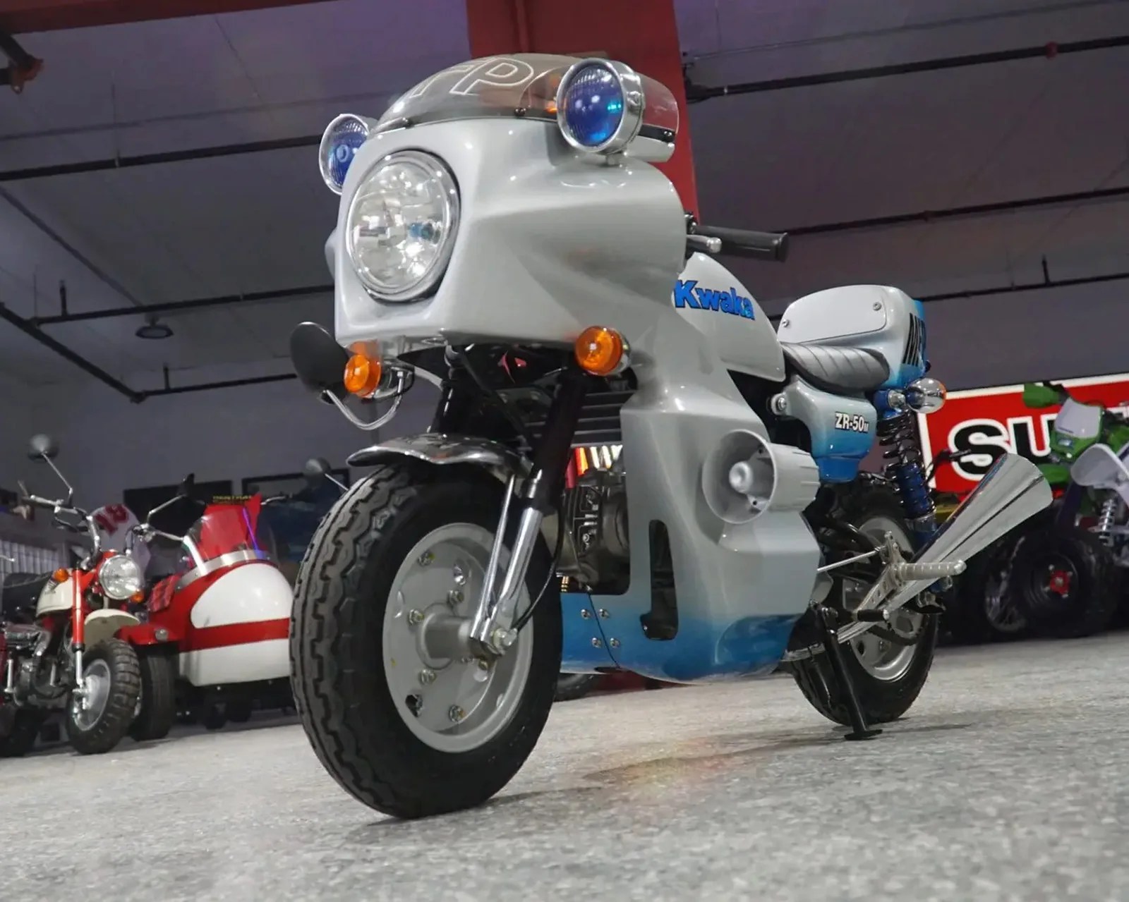 White and blue Kwaka ZR-50 mini motorcycle with round headlights and orange indicators in a showroom.