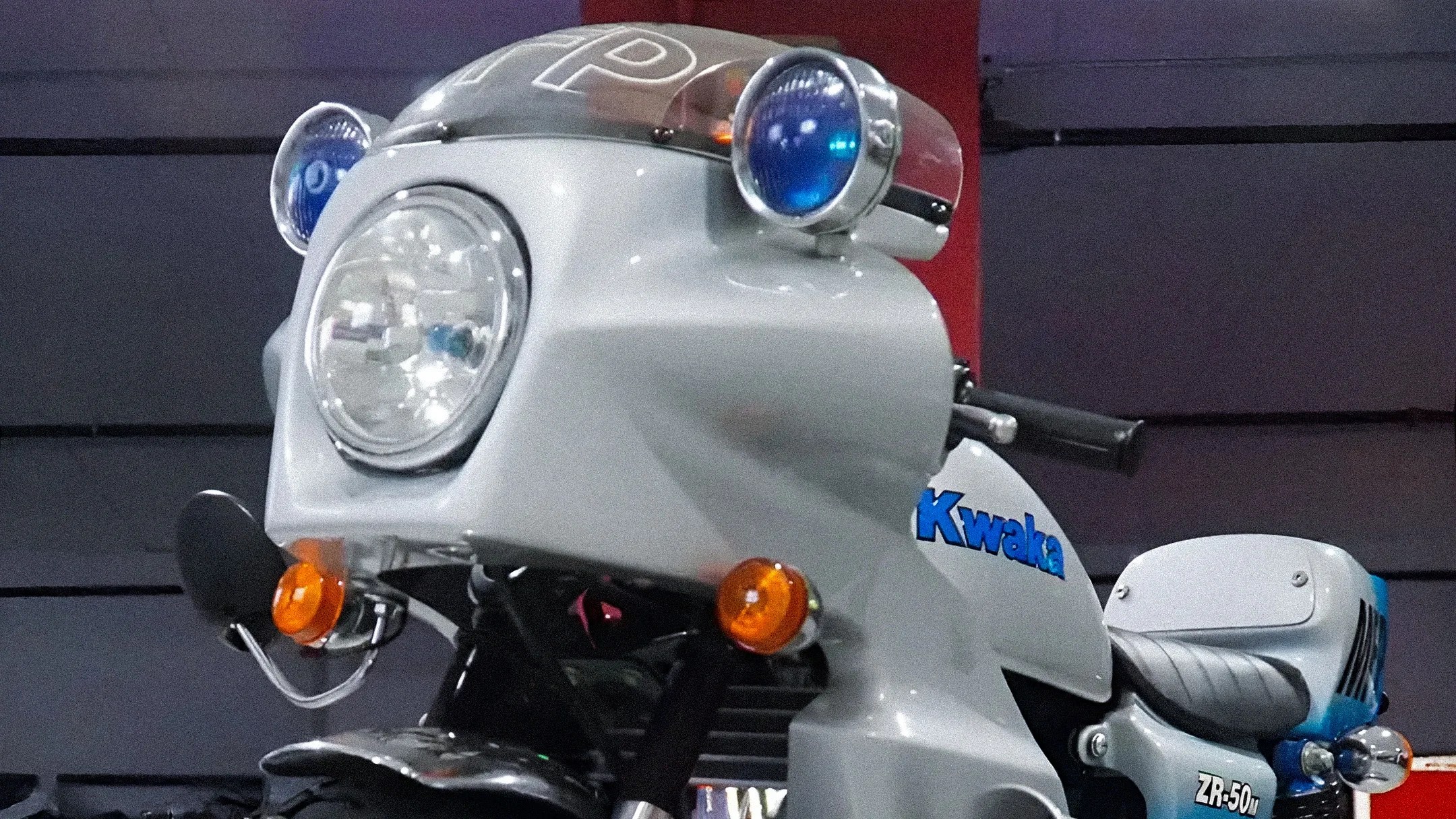 Close-up of a white motorcycle front with large round headlight, blue auxiliary lights, and "Kwaka" and "ZR-50" decals.