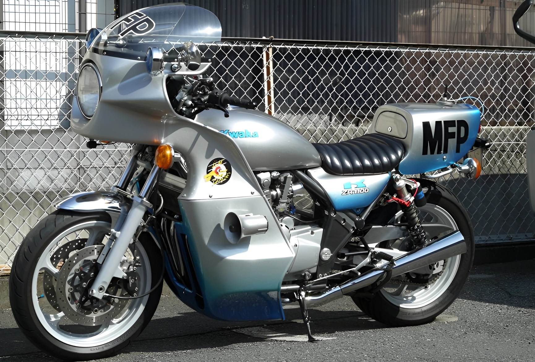 Silver and blue Kawasaki ZR1000 motorcycle with black seat and "MFP" on rear fairing parked on asphalt near chain-link fence.