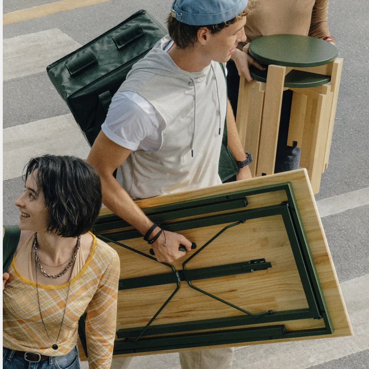 Person carrying a folded wooden table with green metal legs and another person holding stacked wooden stools with green seats.