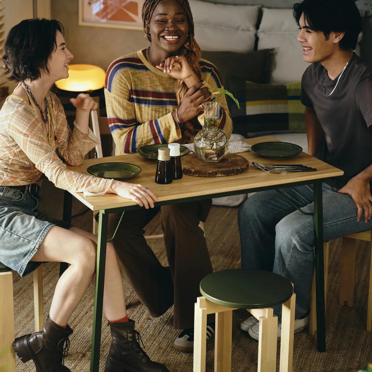 Wooden dining table with green metal legs set with green plates, cutlery, and a glass vase centerpiece with three people sitting around it.