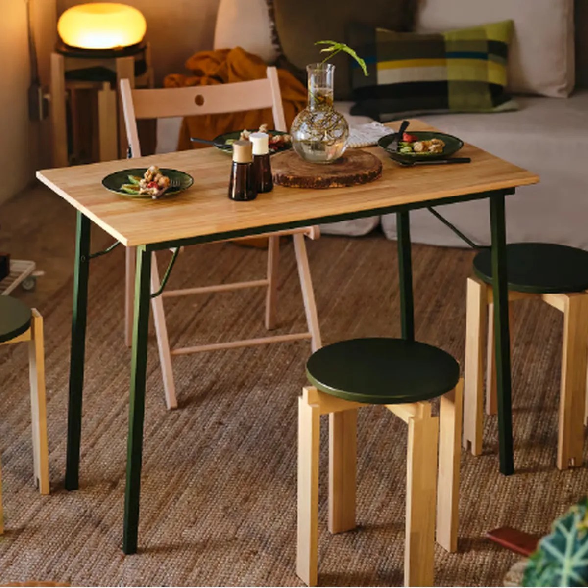 Wooden dining table with black metal legs, set with green plates, salt and pepper shakers, and a glass vase centerpiece.
