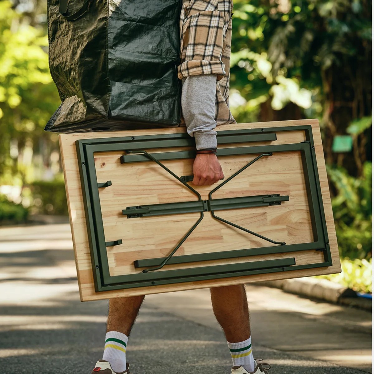 Person holding a folded wooden table with black metal legs and a large black bag over their shoulder.