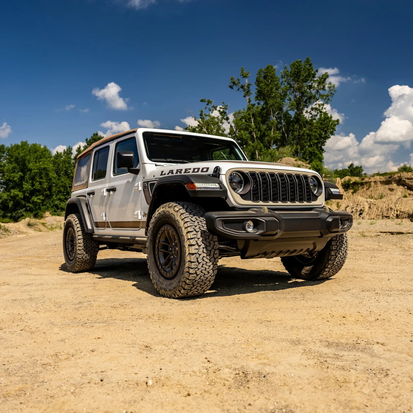 White Jeep Wrangler Laredo with tan soft top and off-road tires parked on dirt with trees and blue sky background.