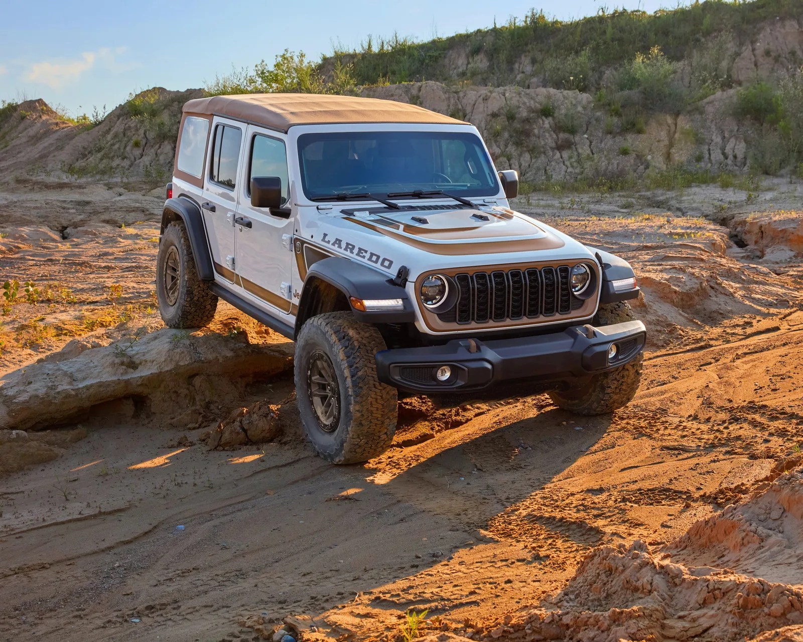 White Jeep Wrangler Laredo with tan soft top parked on rocky, sandy terrain at sunset.