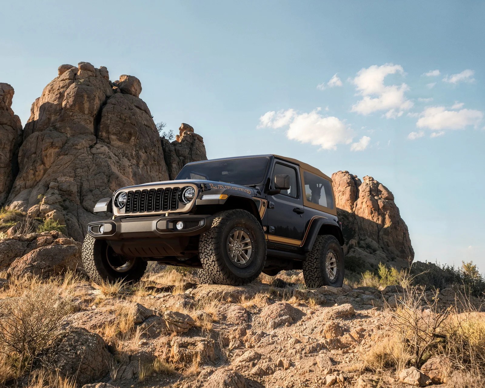 Black Jeep Wrangler with tan roof and accents parked on rocky terrain with large rock formations in the background.