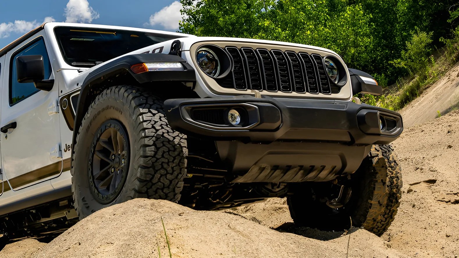 White Jeep Wrangler with black grille and off-road tires climbing a sandy hill with green trees in the background.