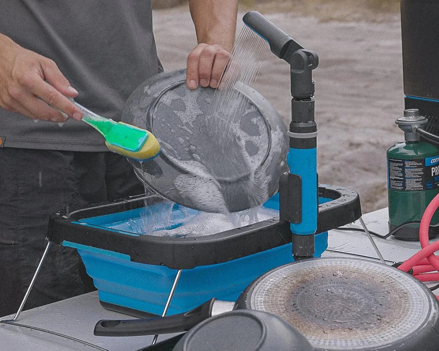 Blue collapsible dishwashing tub with black rim and attached water sprayer being used to wash a pan outdoors.