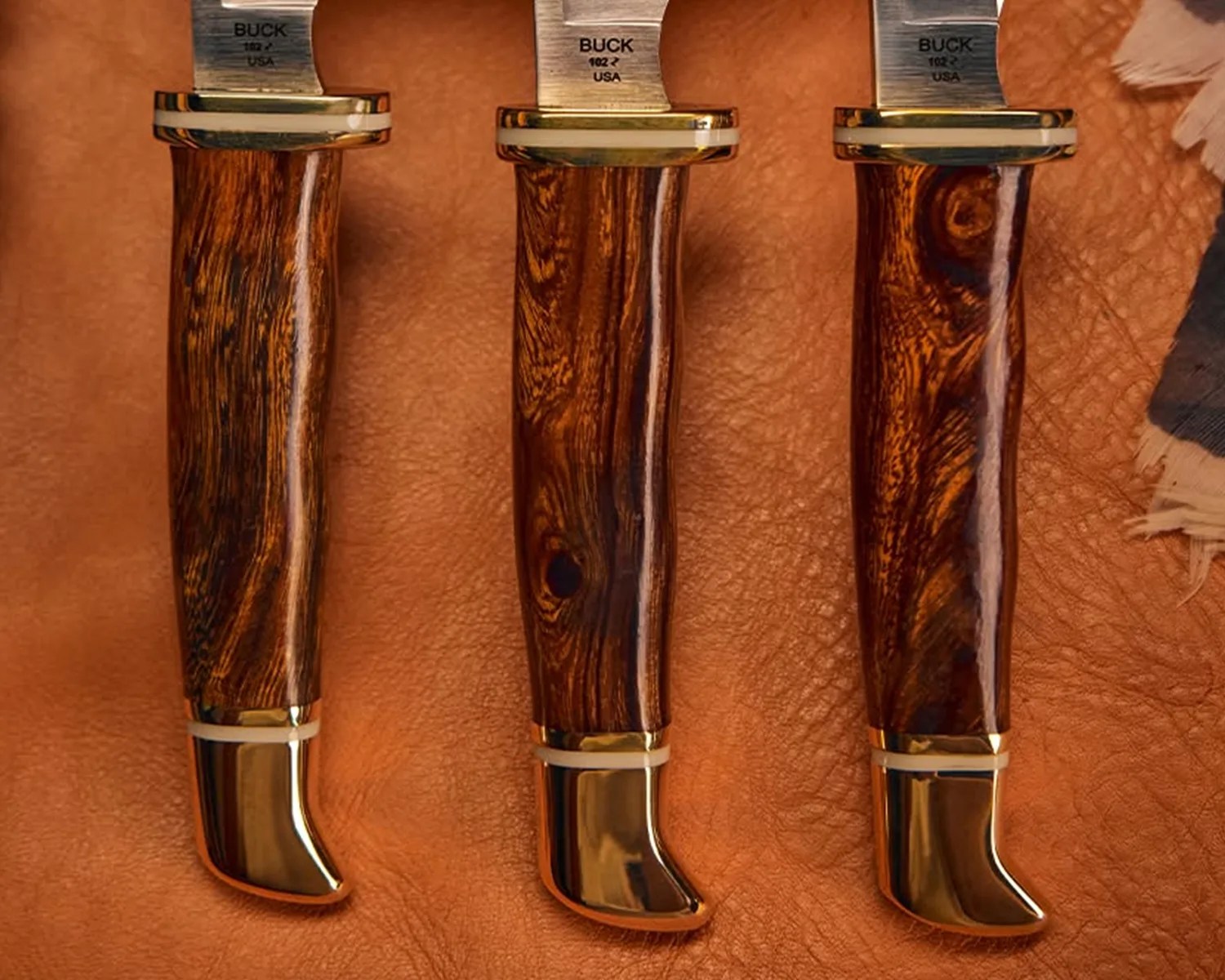 Three Buck 102 USA knives with polished wood handles and brass bolsters on a brown leather surface.
