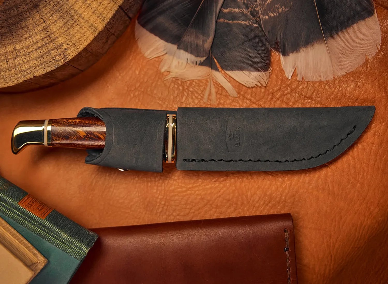 Hunting knife with wooden handle and brass pommel in a black leather sheath on a brown leather surface.
