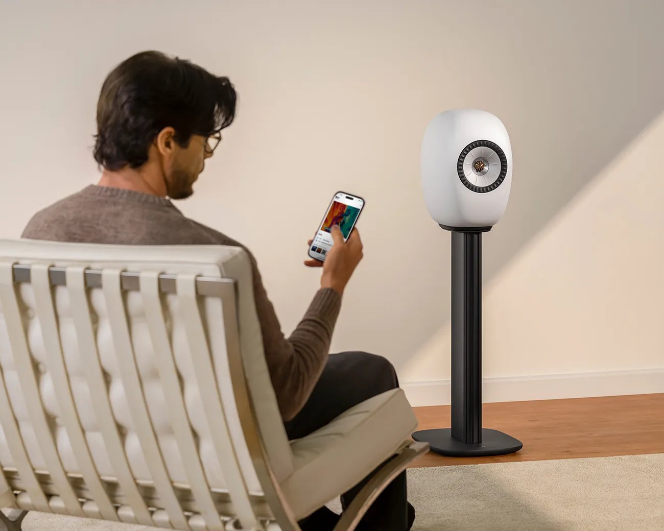 White oval speaker on a black stand next to a person sitting on a white cushioned chair holding a smartphone.